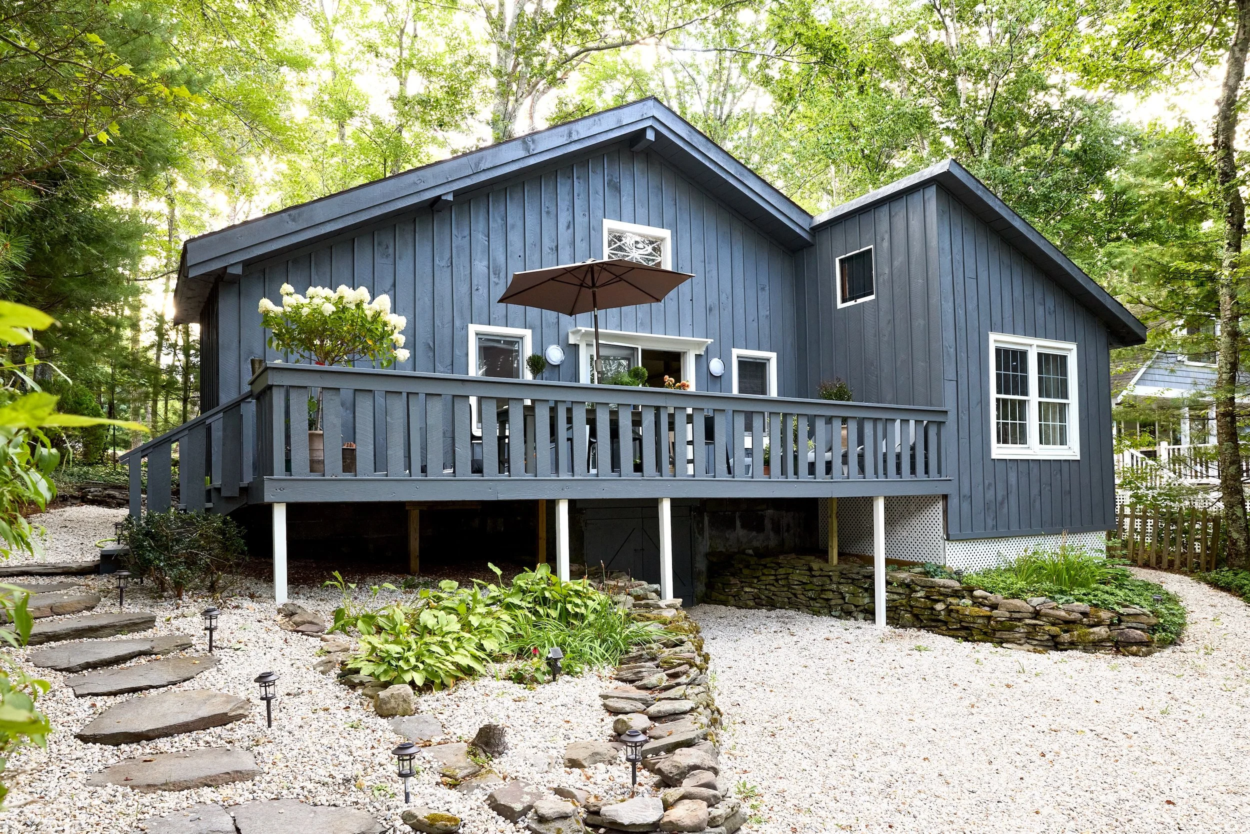 A blue wooden house with a raised porch, surrounded by trees and a gravel yard with stone steps and landscaping.