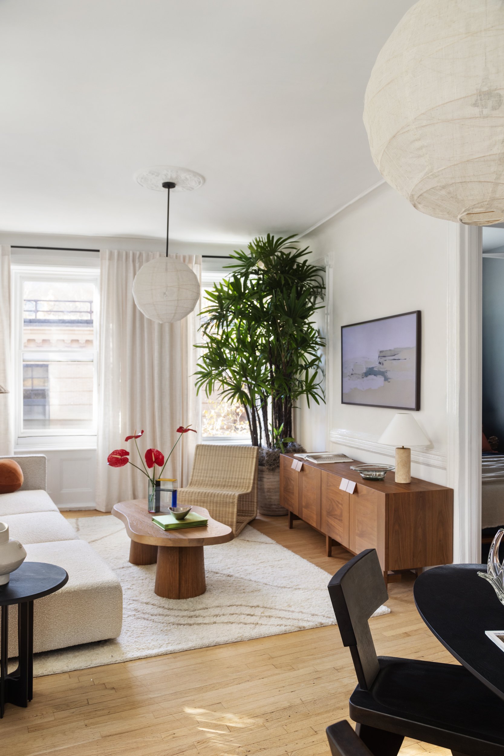Living room with beige sofa, wooden coffee table, black side table, woven chair, indoor plant, white curtains, hanging white paper lanterns, wall-mounted artwork, and wooden sideboard with a table lamp.