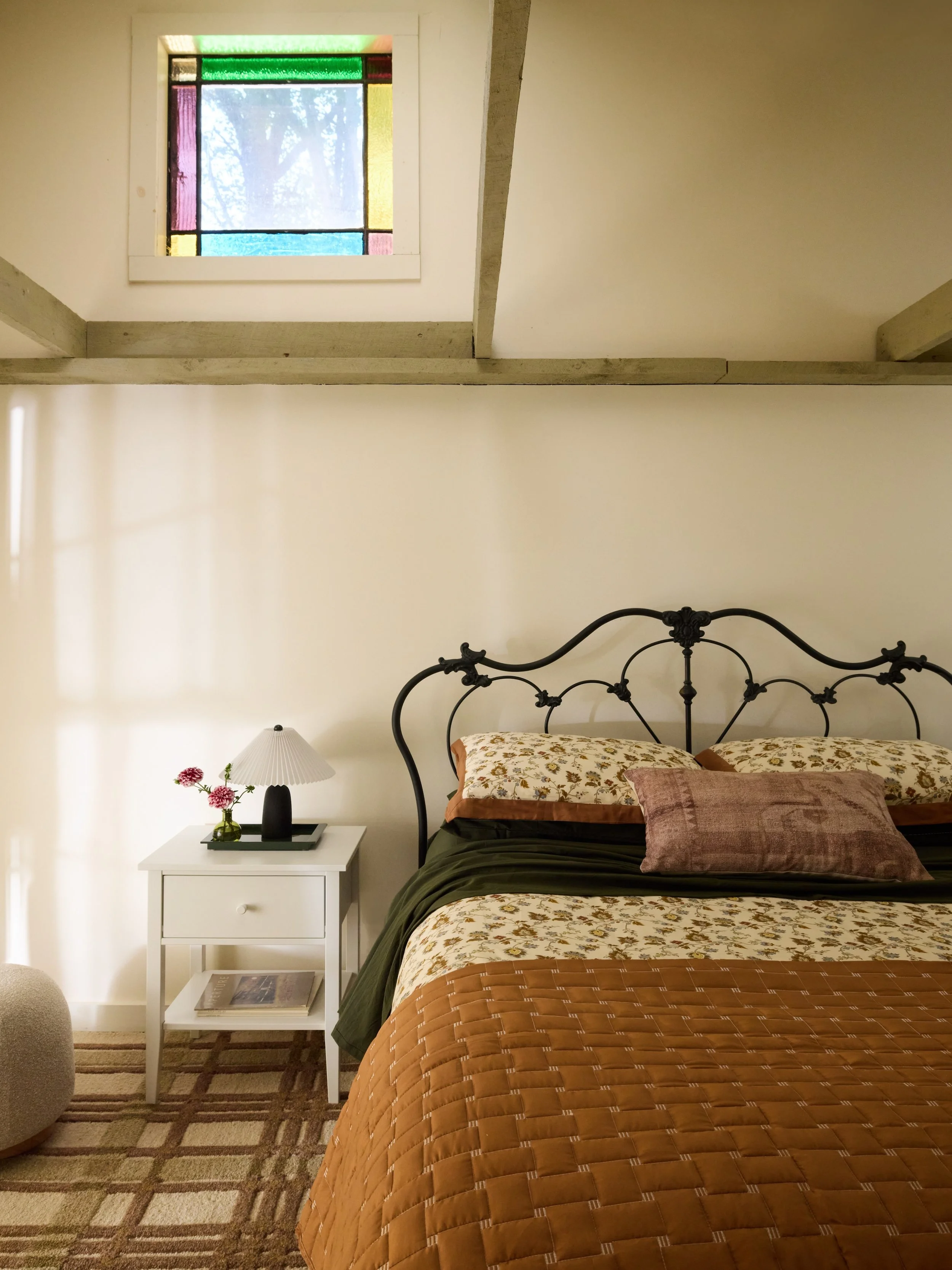 A cozy bedroom with a black iron bed frame, floral-patterned bedding, and pillows, a white nightstand with flowers and lamp, and a stained glass window in the ceiling.