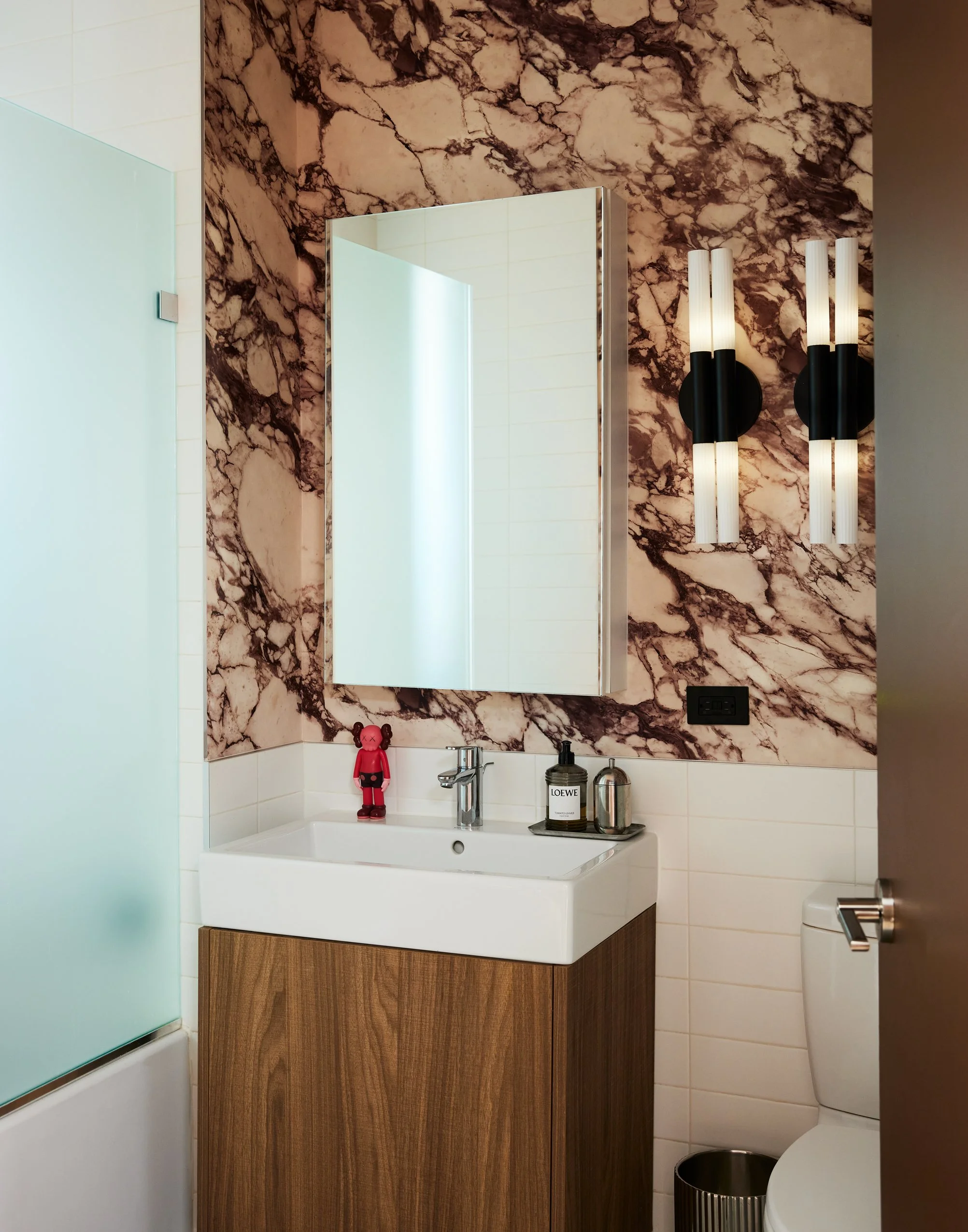 Bathroom with marble wall, mirror, white sink, wooden cabinet, modern black light fixtures, and a toilet.