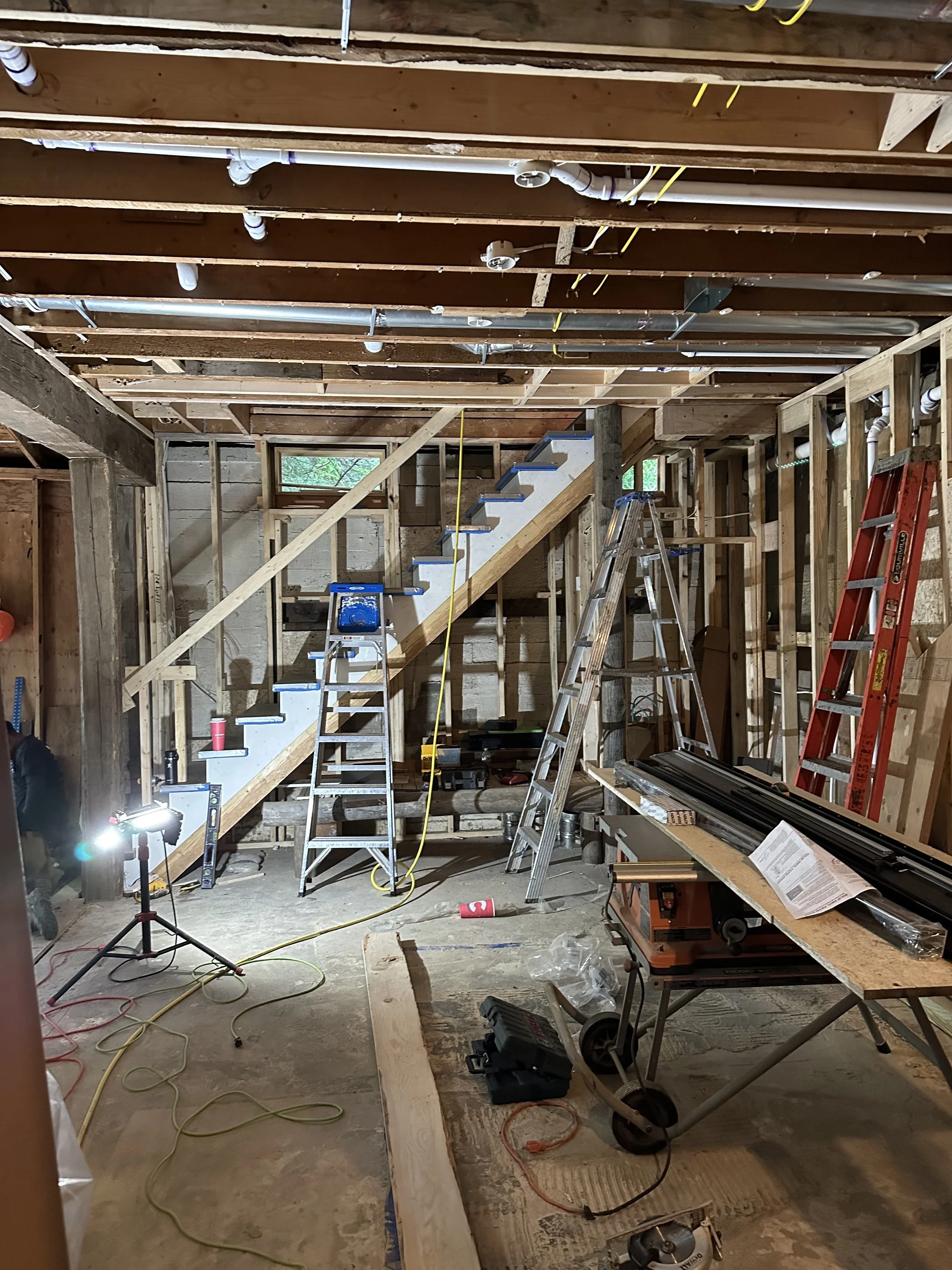 Interior view of a house under construction with exposed wooden framing, stairs, ladders, construction tools, and electrical wiring.