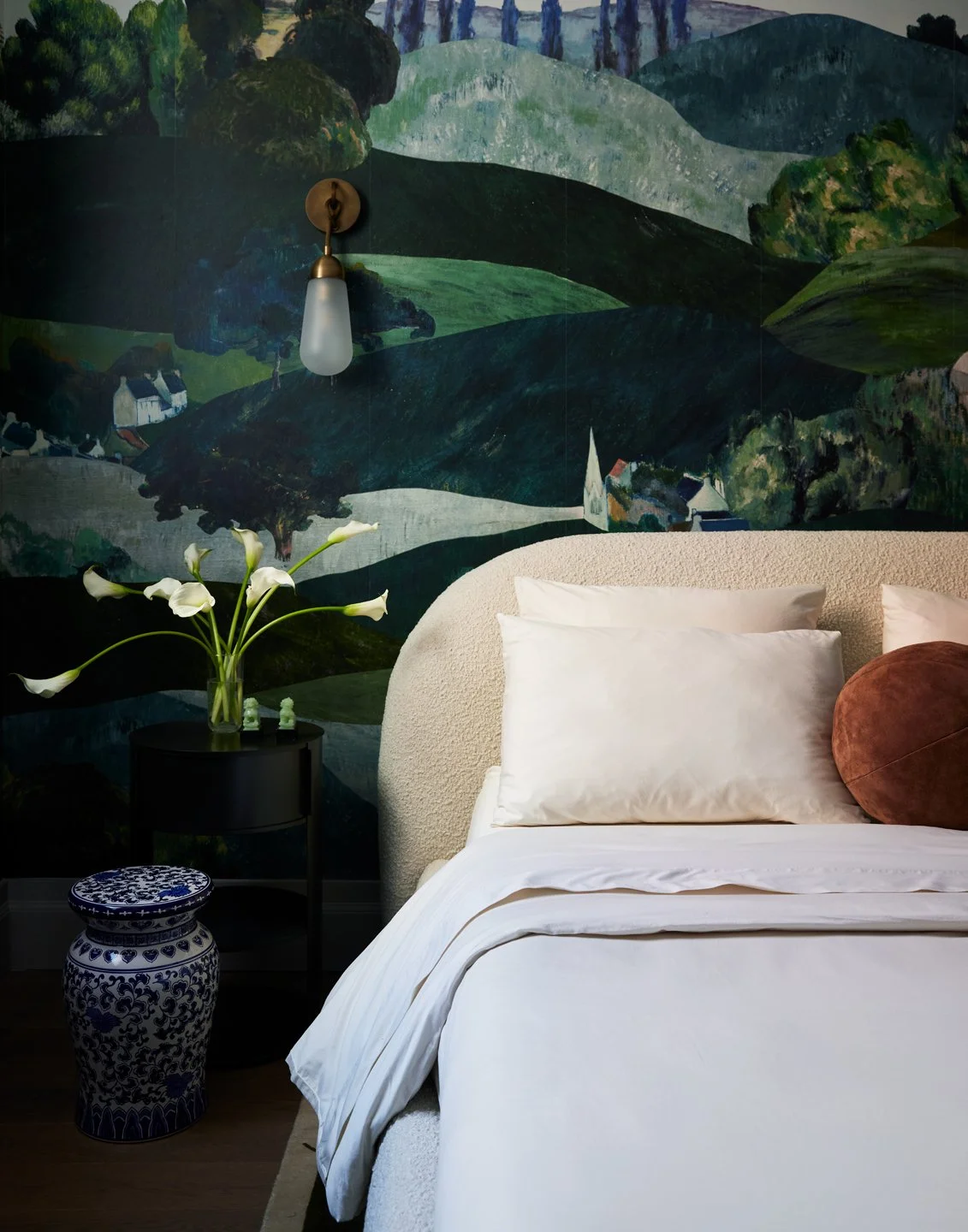 A bedroom with a white bed featuring pillows and a rounded headboard, a black side table with a vase of white calla lilies, a decorative blue and white ceramic stool, and a mural of a green landscape with trees and houses on the wall. A wall-mounted light fixture is also visible.
