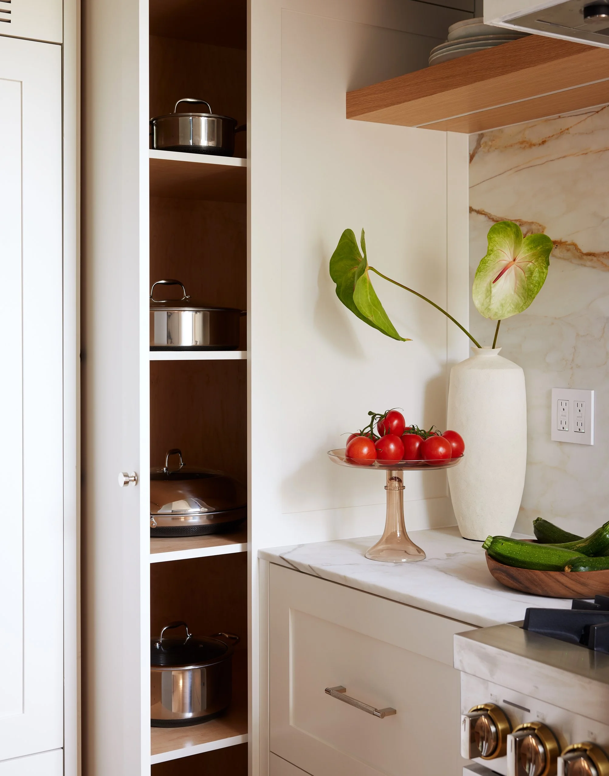 Kitchen countertop with a glass cake stand holding cherry tomatoes, a white vase with green leaves, a wooden bowl with zucchinis, and a stove with gold knobs. Open cabinet with stainless steel pots and a marble backsplash.