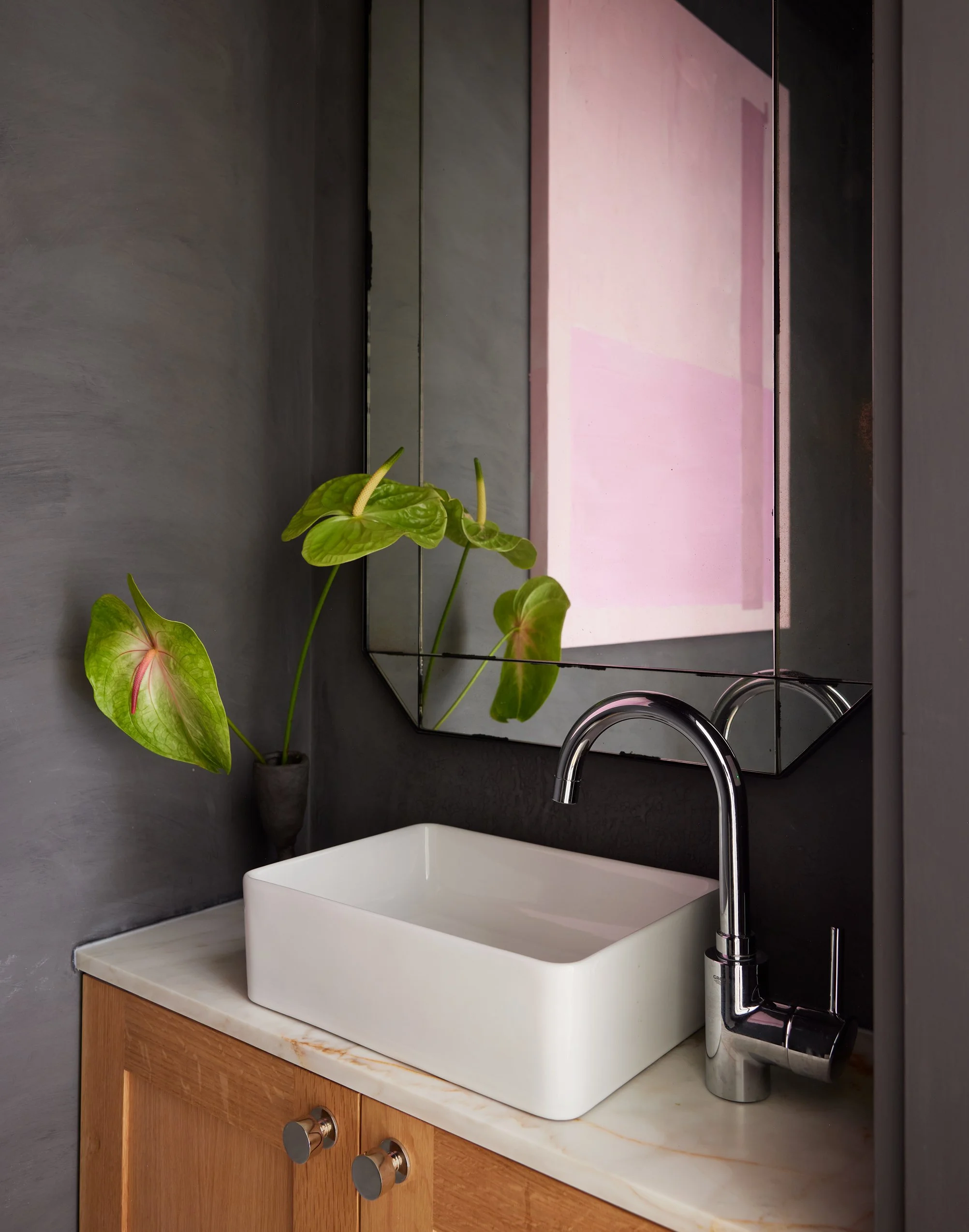 Bathroom sink area with a square white vessel sink, a modern chrome faucet, a frameless mirror, dark gray walls, a wooden cabinet with metallic knobs, and a potted plant with large green leaves.