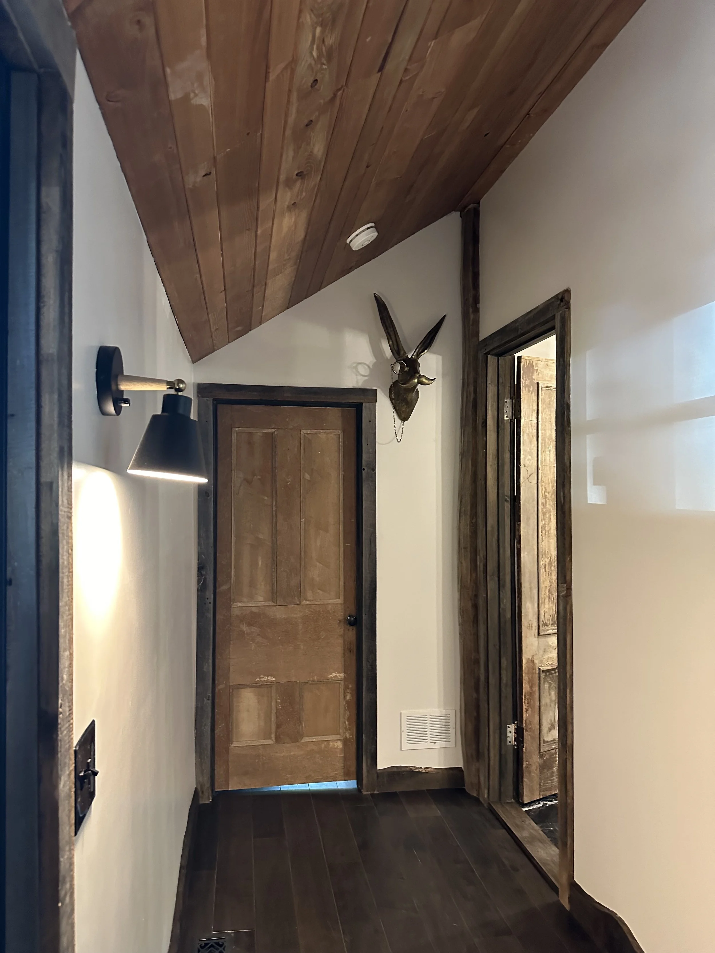 Interior view of a rustic-style hallway with glossy dark wood flooring, wooden doors framed in distressed wood, a sloped wooden ceiling, and a mounted decorative animal head with antlers.