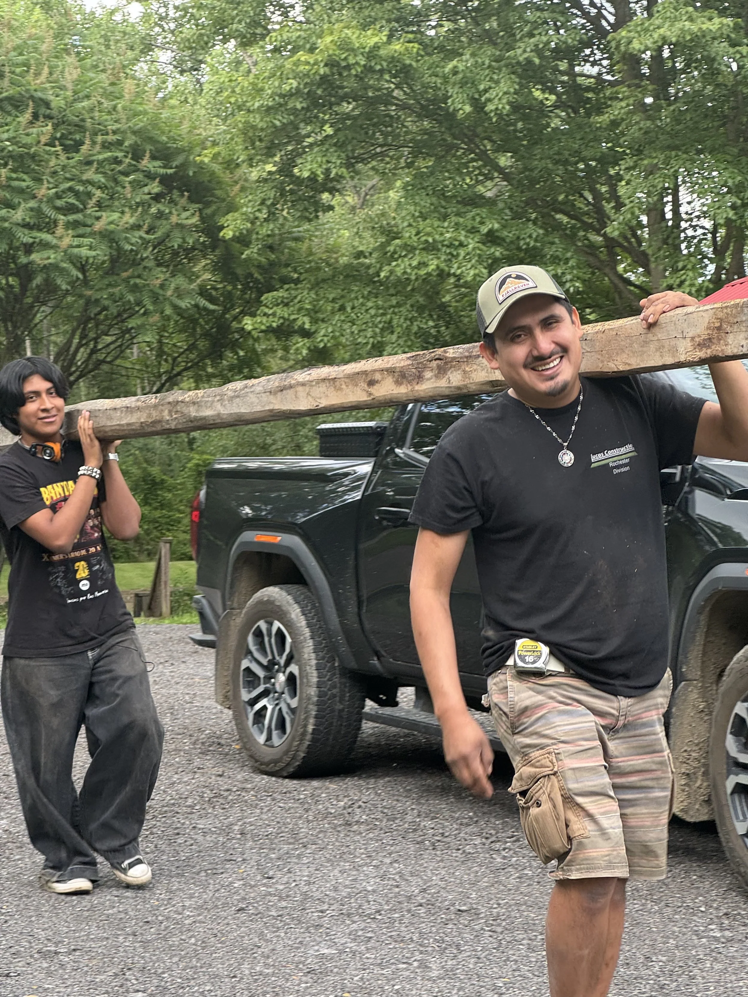 Two men carrying a large wooden beam in an outdoor setting with trees and a black vehicle in the background.