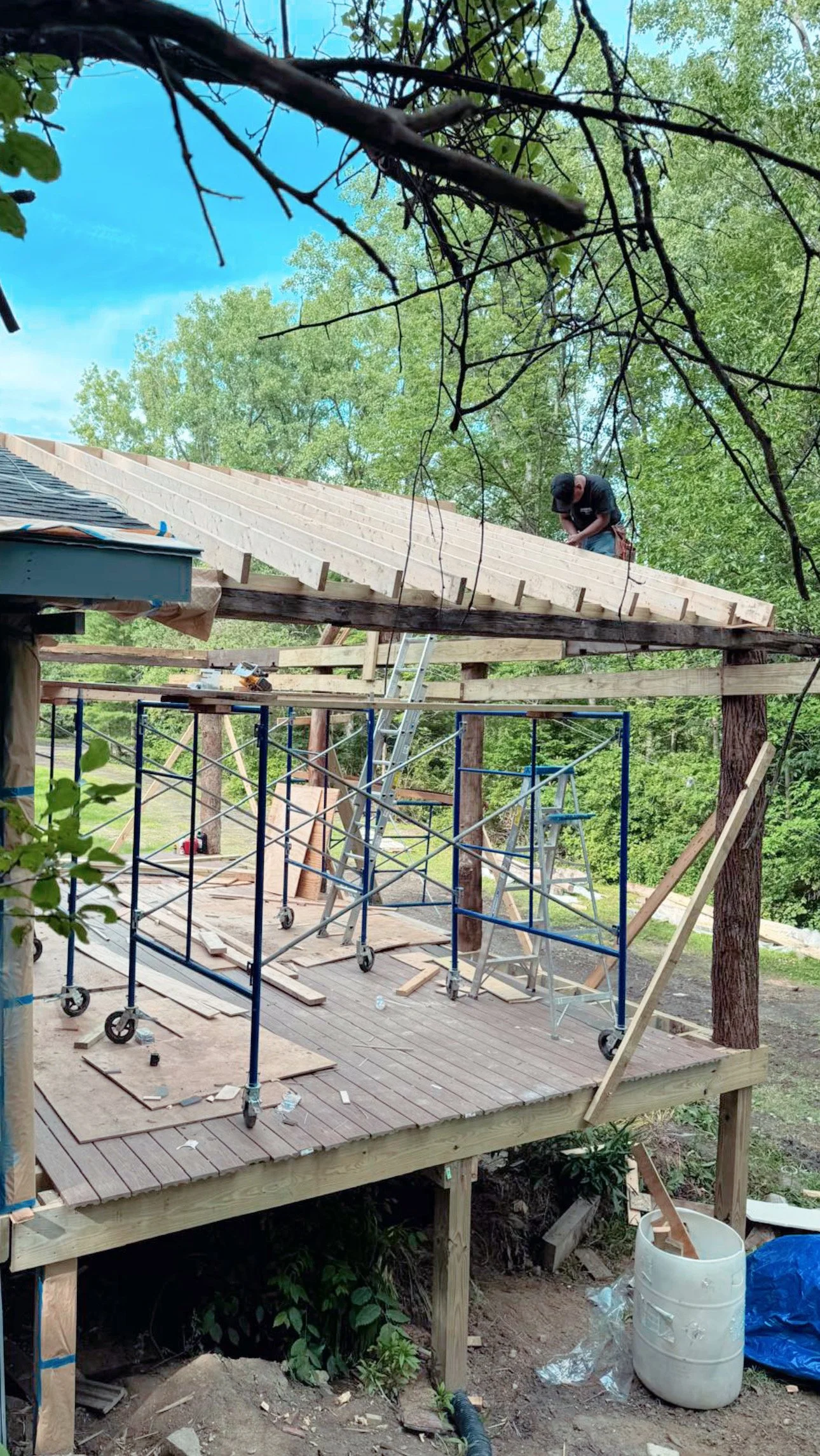A person working on roof construction using scaffolding and ladders, surrounded by trees and construction tools.