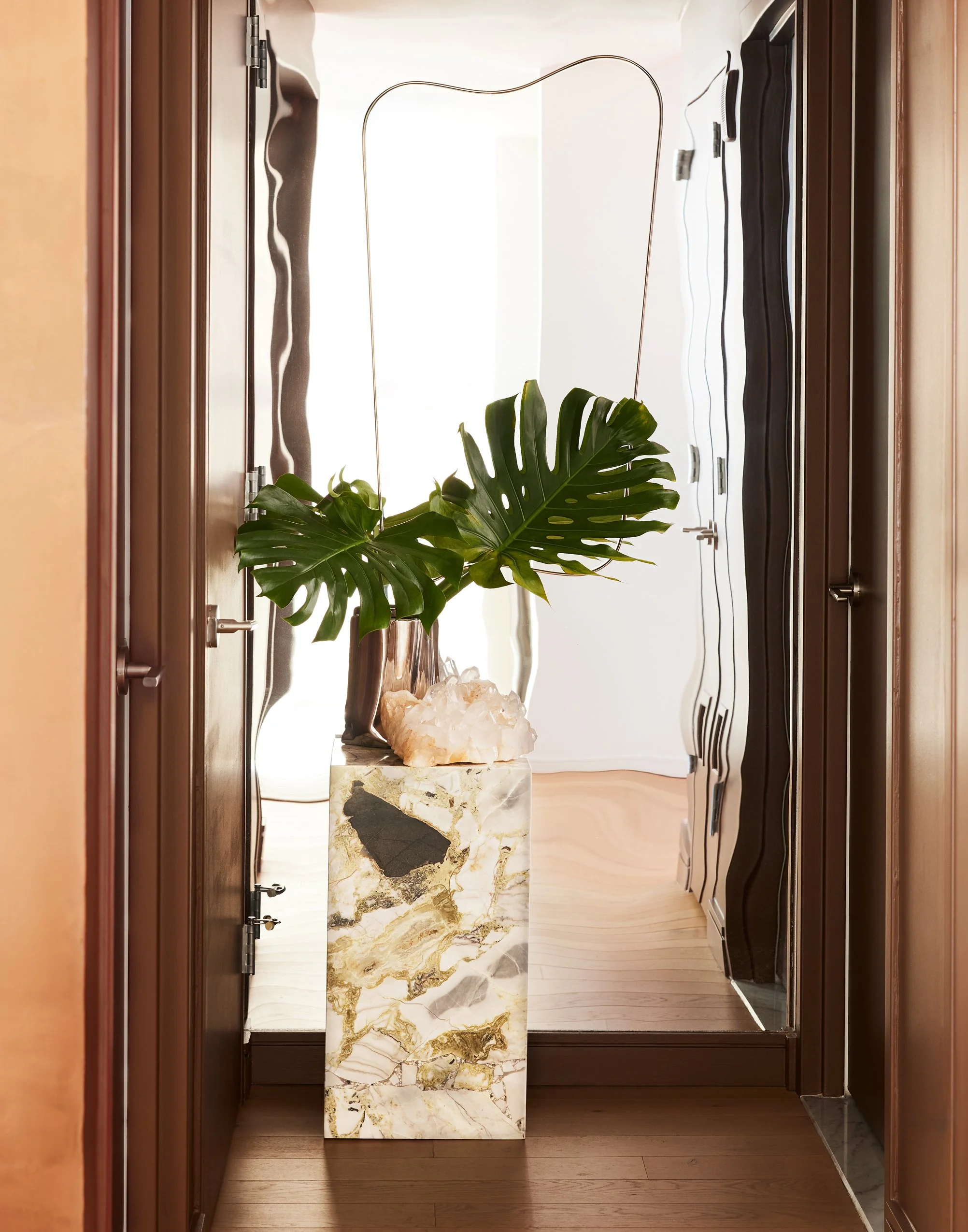 A tall marble pedestal with gold, black, and gray veining, holding a large Monstera plant with broad, split green leaves. Behind it, a mirror reflects the room, and a crystal-like decorative object rests on the pedestal. The setting appears to be a hallway or entryway with wood flooring and dark wood door frames.