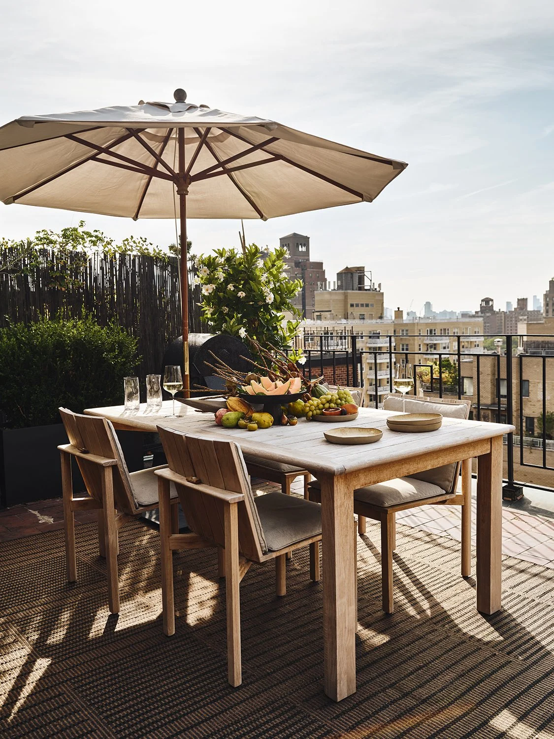 Outdoor rooftop patio with a wooden dining table, chairs, a large beige umbrella, a fruit centerpiece, and a city skyline background.