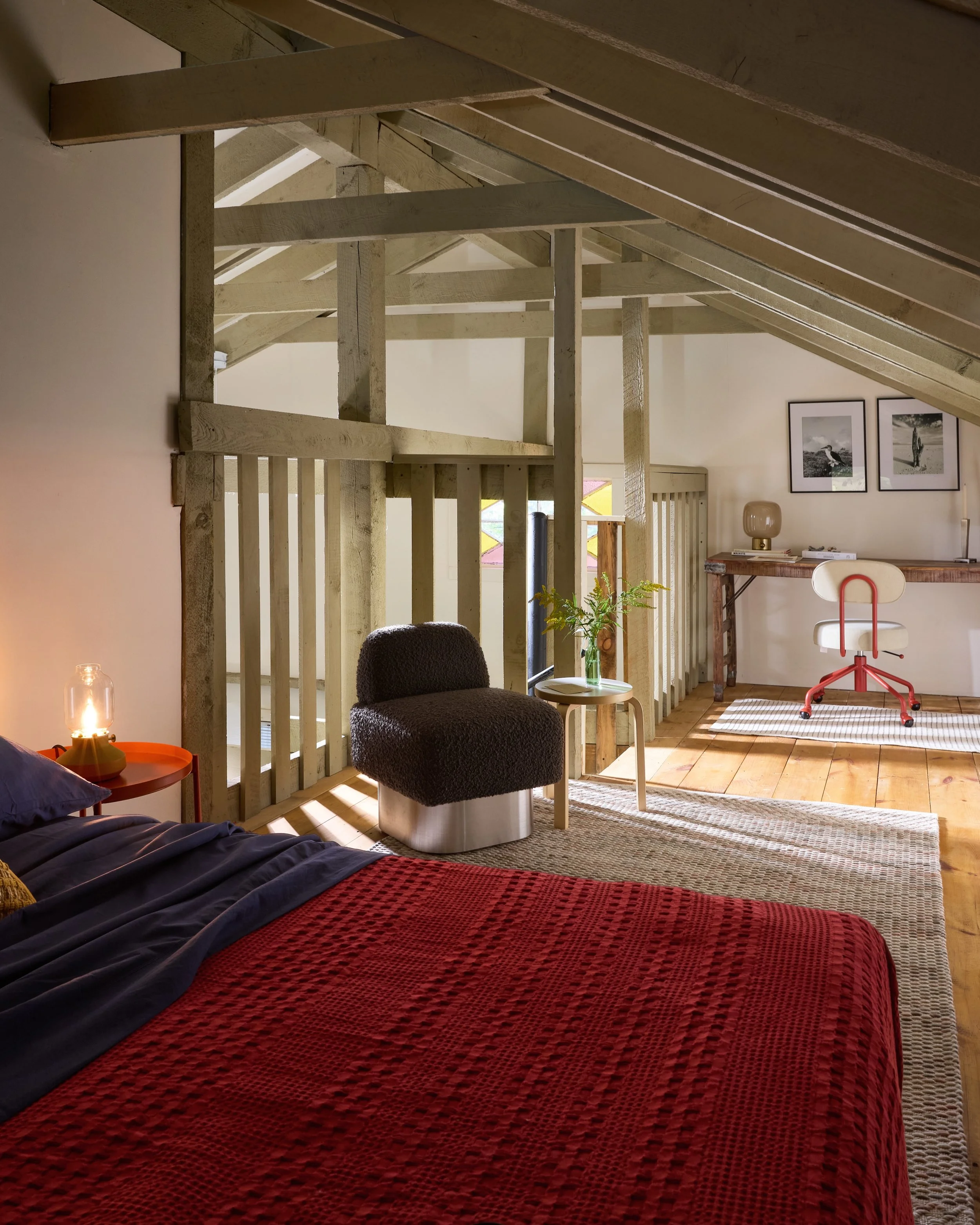 A cozy bedroom with a bed covered in a red textured blanket, a nightstand with a lantern, a plush dark gray armchair, a small side table with green foliage, a rustic-style desk in the background with framed black-and-white photos on the wall, and a white office chair with red accents on a striped rug. The room features exposed wooden beams and natural light.