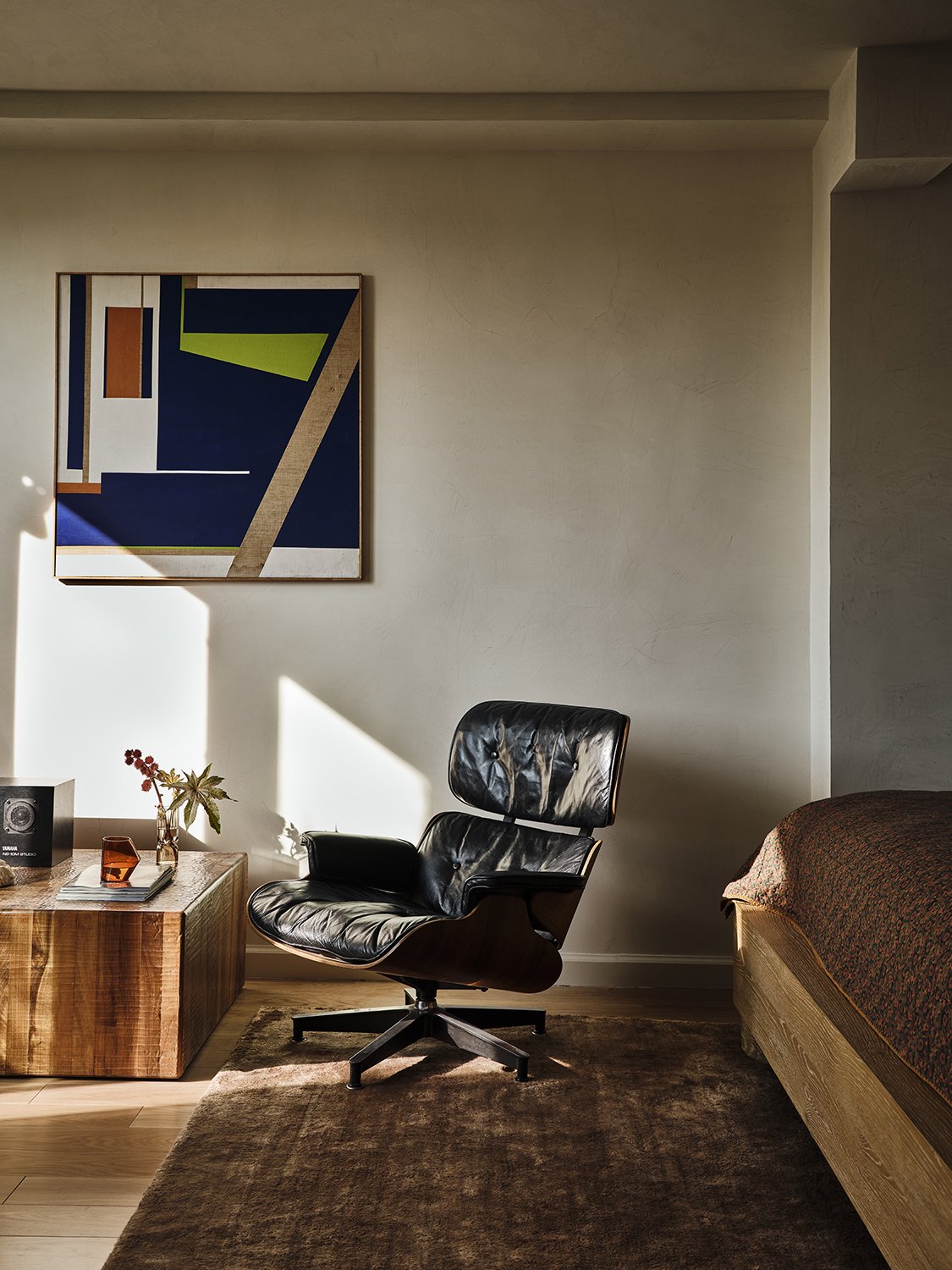 A living room corner with a modern black leather chair, a wooden side table with a small plant, a book, an orange glass, and a speaker. There is a colorful abstract painting on the wall, and sunlight casting shadows on the wall and floor. A bed with a patterned blanket is partially visible on the right.