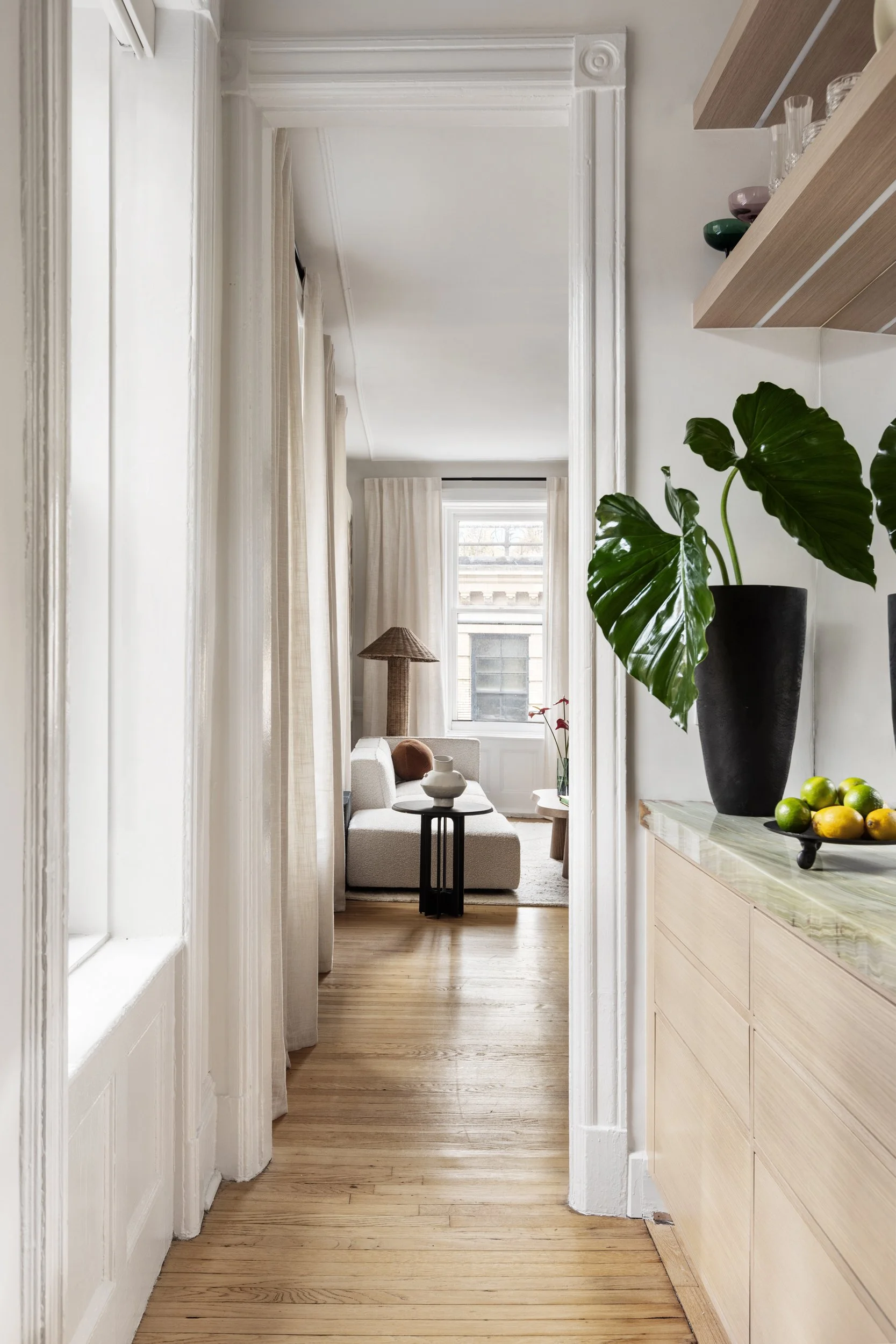 View through a doorway into a living room with a white sofa, round black side table, window with white curtains, and a floor lamp with a woven lampshade. There is a large green plant and a bowl of lemons on a wooden cabinet in the foreground.