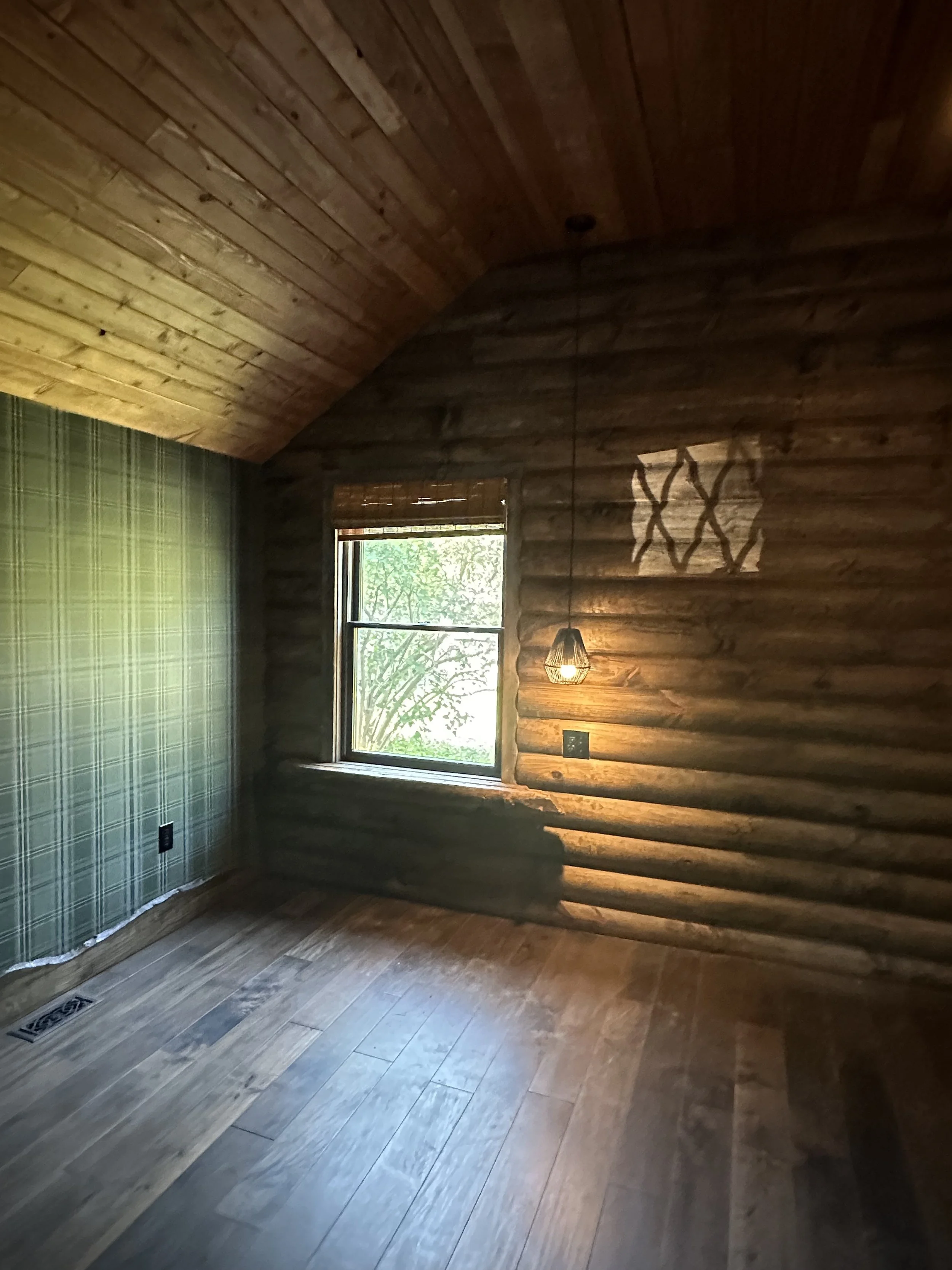 A cozy room with wooden logs wall, a window with bamboo shade, green checkered wallpaper on one wall, a hanging pendant light, and wooden flooring. Sunlight streams through the window.
