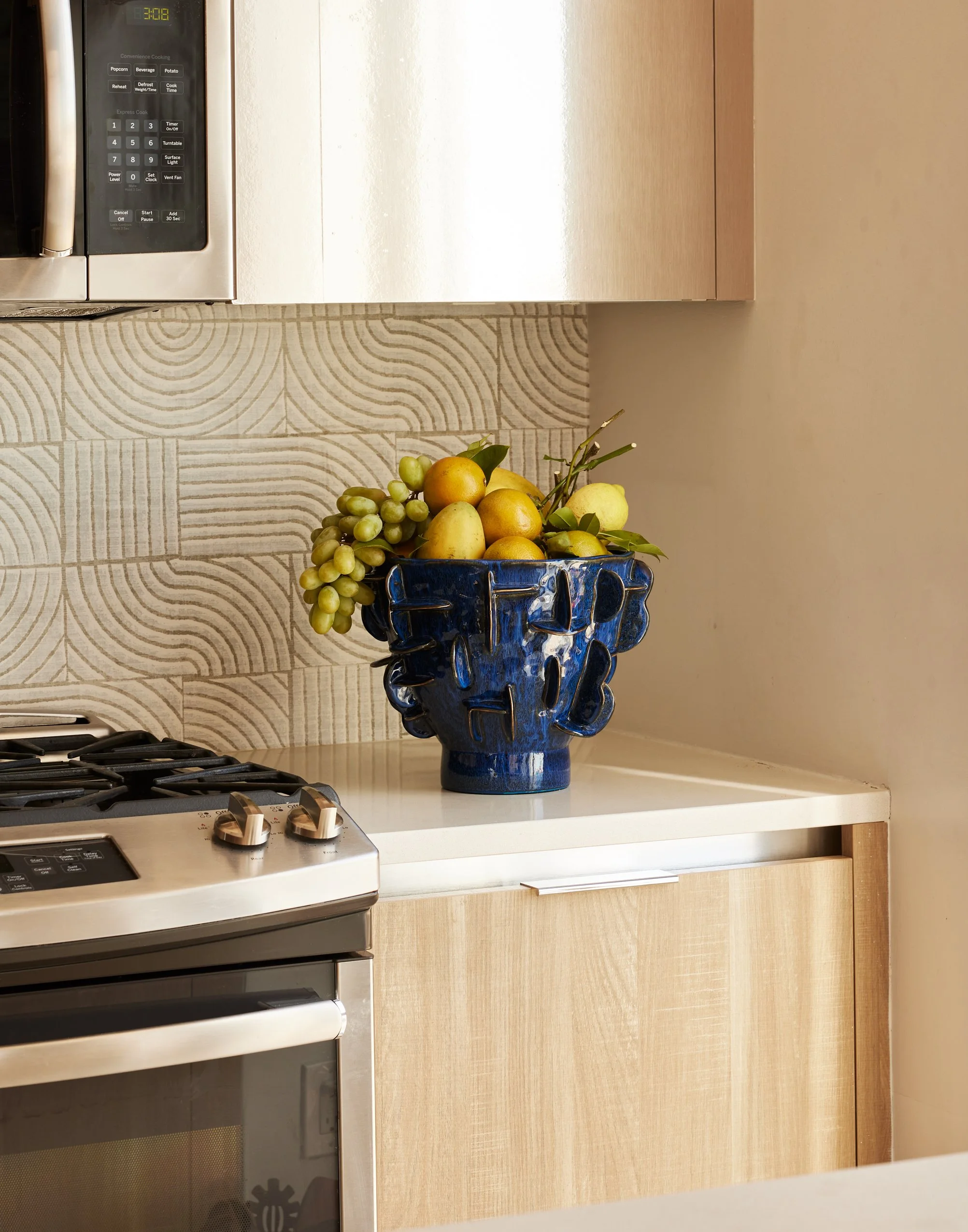A blue ceramic vase filled with various fruits, including grapes, pears, and citrus, placed on a white kitchen countertop next to a stove and a wooden cabinet.
