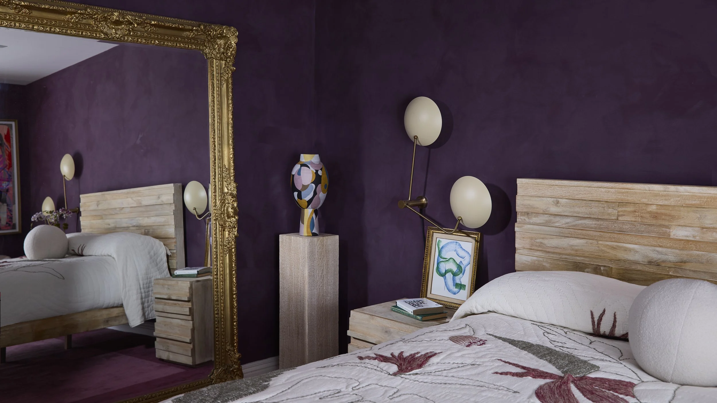 A bedroom with purple walls, light wood headboards, and decorative lamps. A large ornate gold mirror, a colorful vase on a pedestal, and framed artwork on the bedside table are visible.