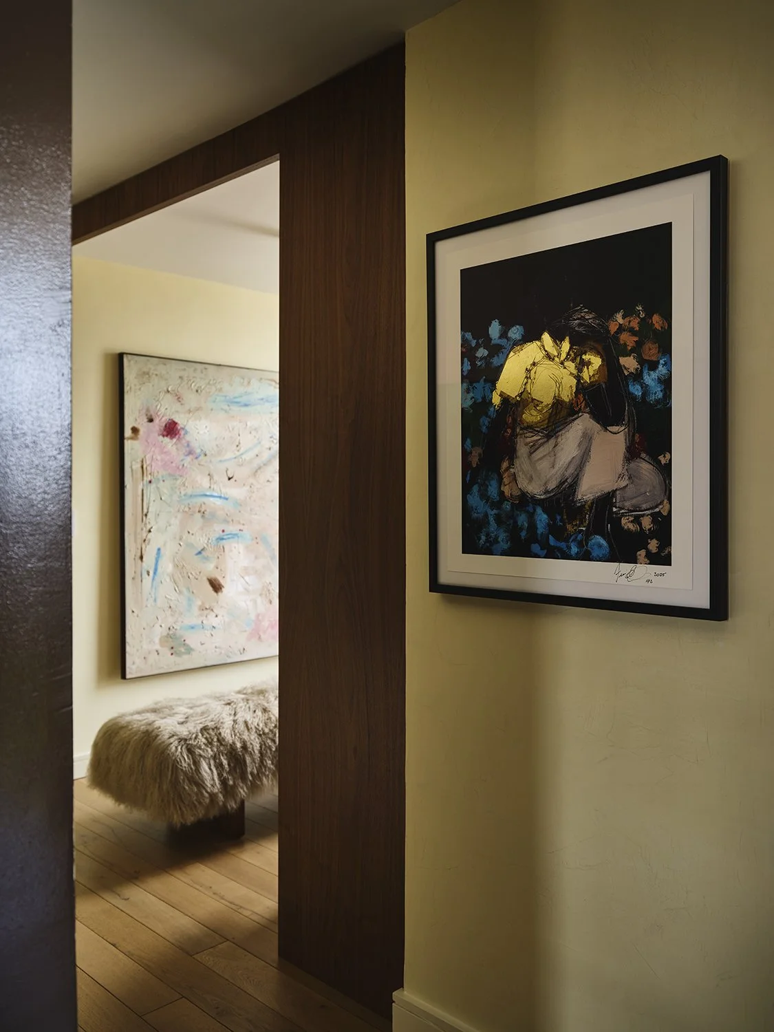 Interior view of a corner in an art gallery or home, showing a framed painting of a person sitting with flowers in the background on a cream-colored wall, a second abstract painting on a yellow wall in the background, a wooden panel, a fluffy ottoman, and hardwood flooring.