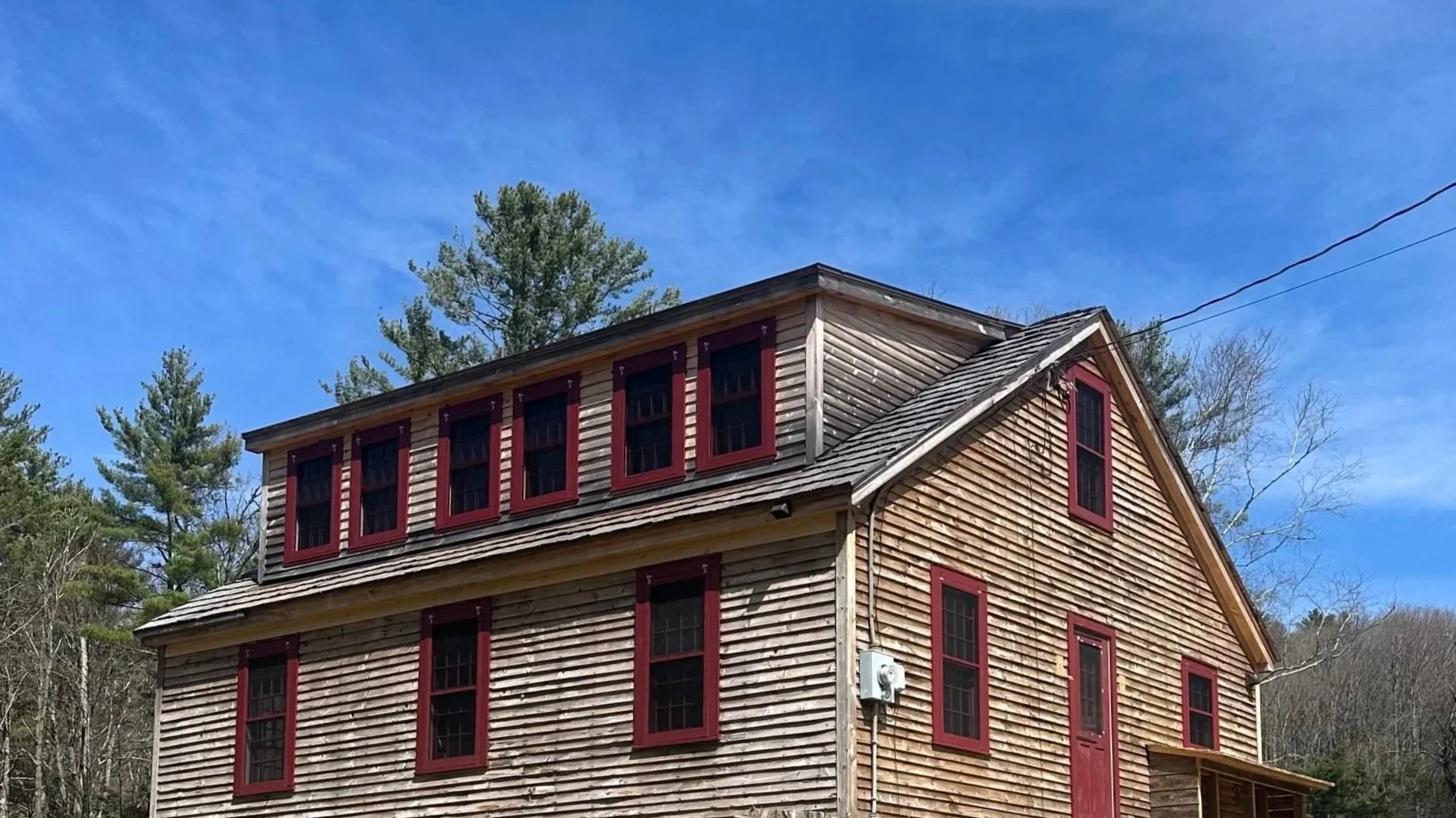 A two-story wooden house with a pitched roof, multiple windows with red trim, and surrounded by trees against a blue sky.