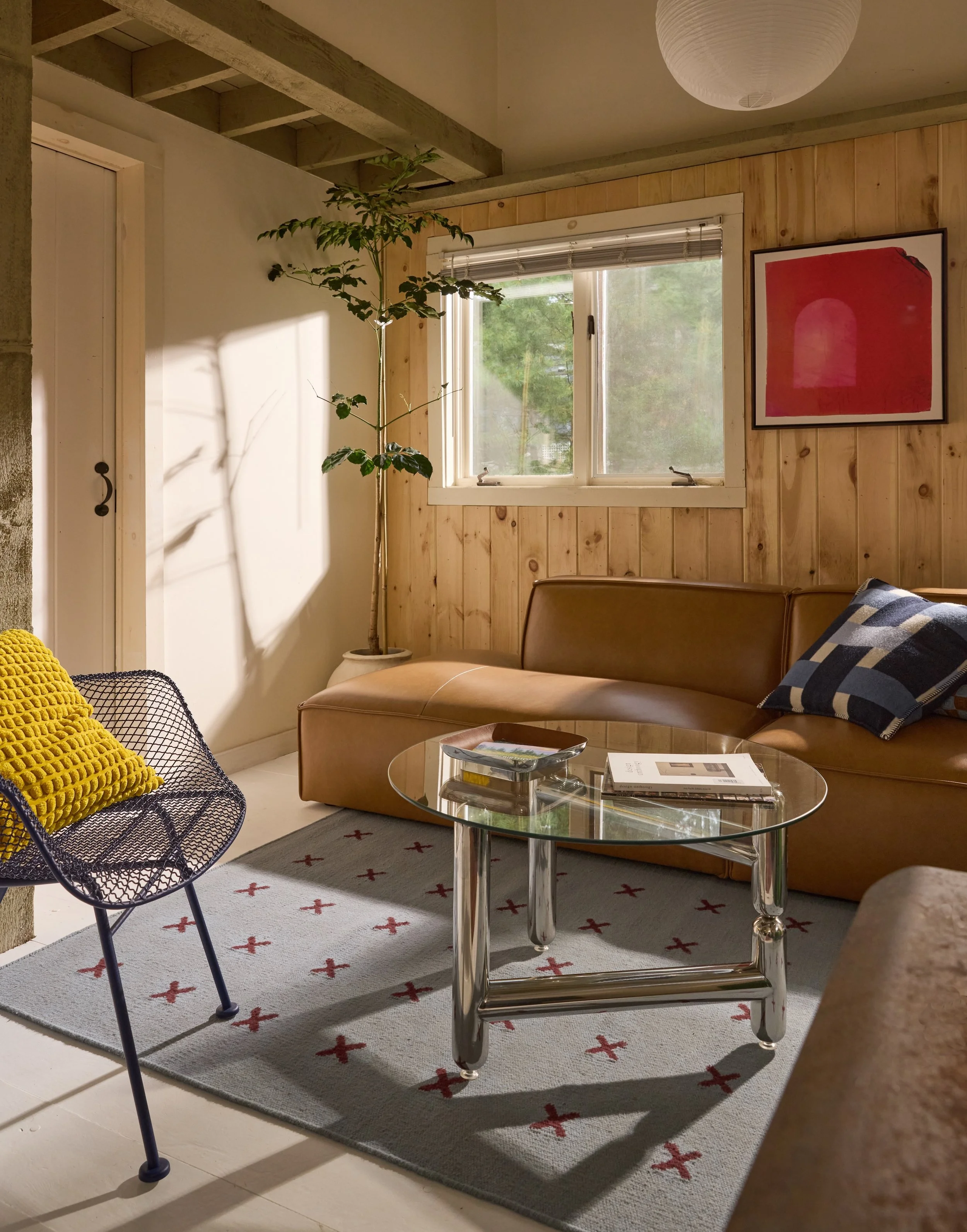 A cozy living room with a brown leather sofa, a glass coffee table, a patterned rug, a black wire chair with a yellow pillow, a tall potted plant, a window with blinds, and a framed red artwork on wood-paneled wall.