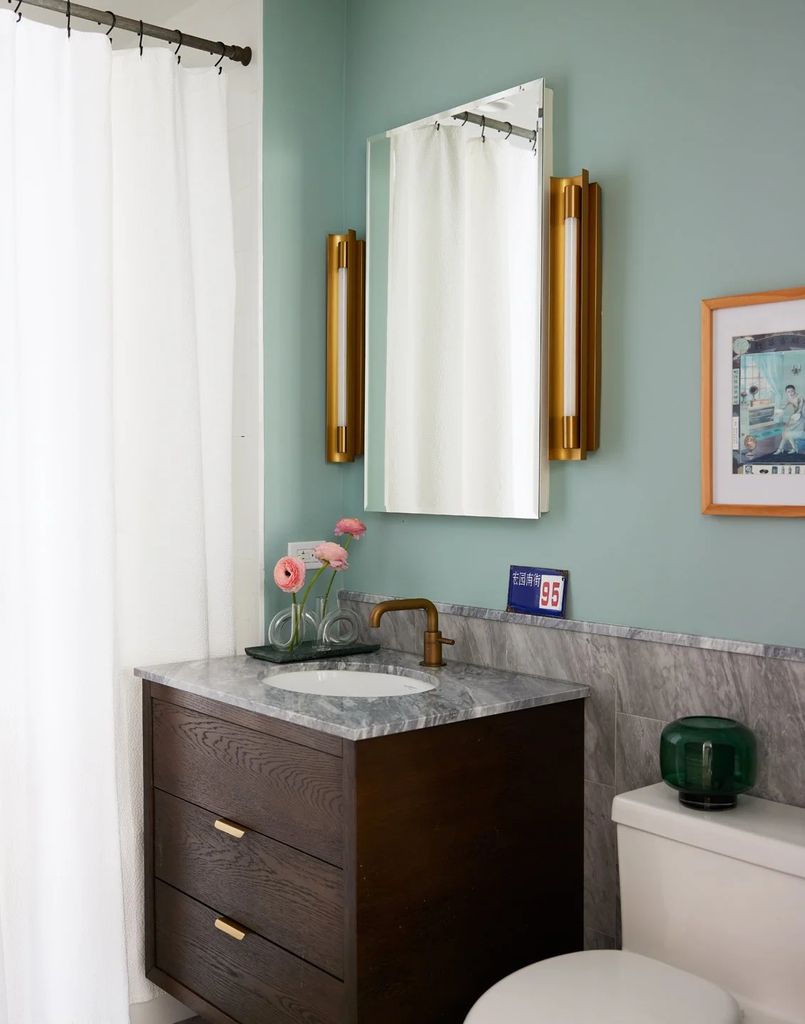 Modern bathroom with a dark wooden vanity, gray marble countertop, and a round white sink. Above, a mirror is flanked by gold vertical wall sconces. A small vase with pink flowers is on the countertop. To the right, part of a white toilet with a green diffuser on top, beige tile wainscoting, and light blue-green painted walls are visible. White curtains hang on a black rod on the left side.
