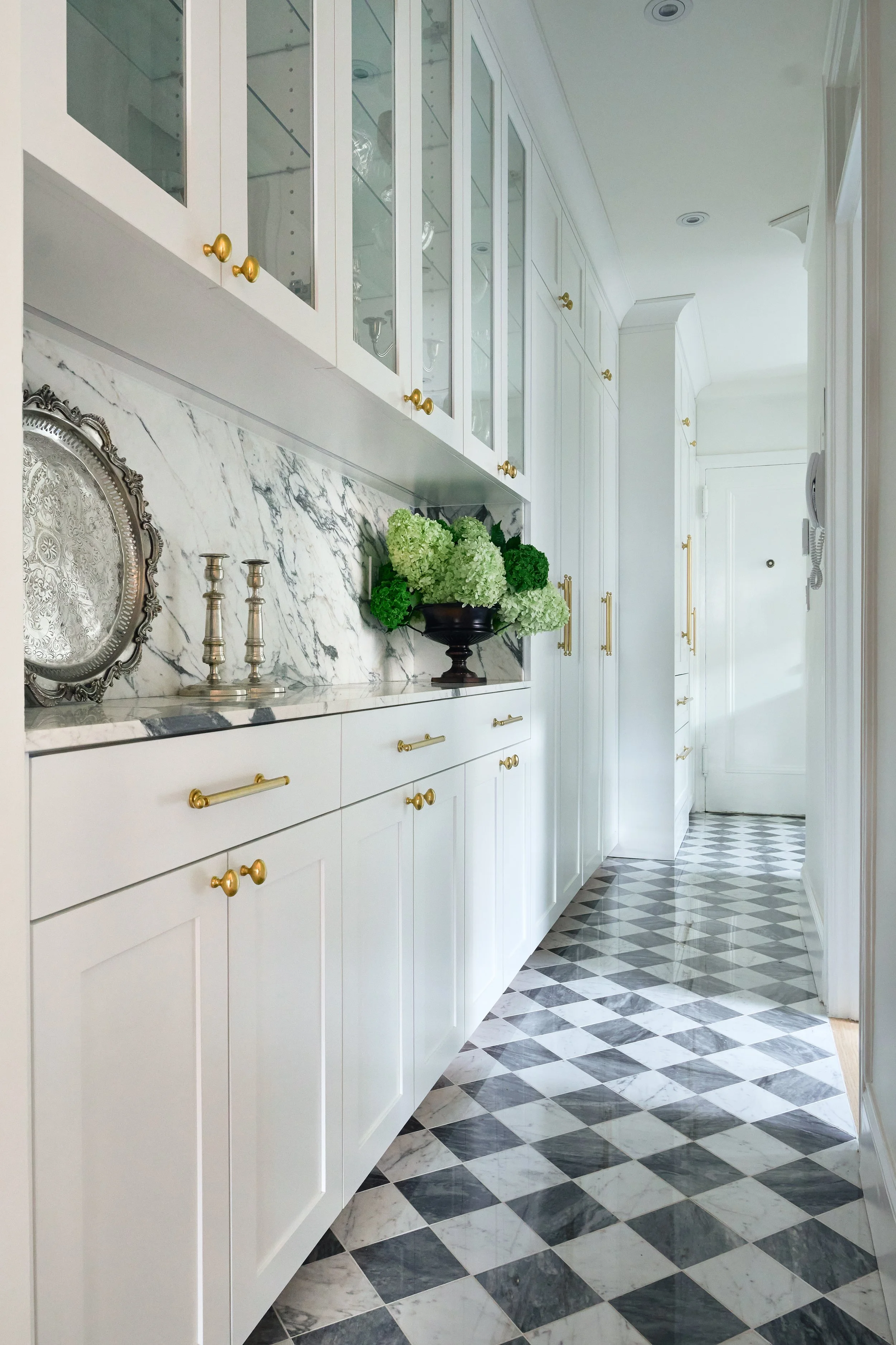 A hallway with white cabinets, glass-front upper cabinets, and a marble backsplash, decorated with silver candlesticks, a silver tray, and a black vase with white and green flowers. The floor has a black and white checkered pattern.