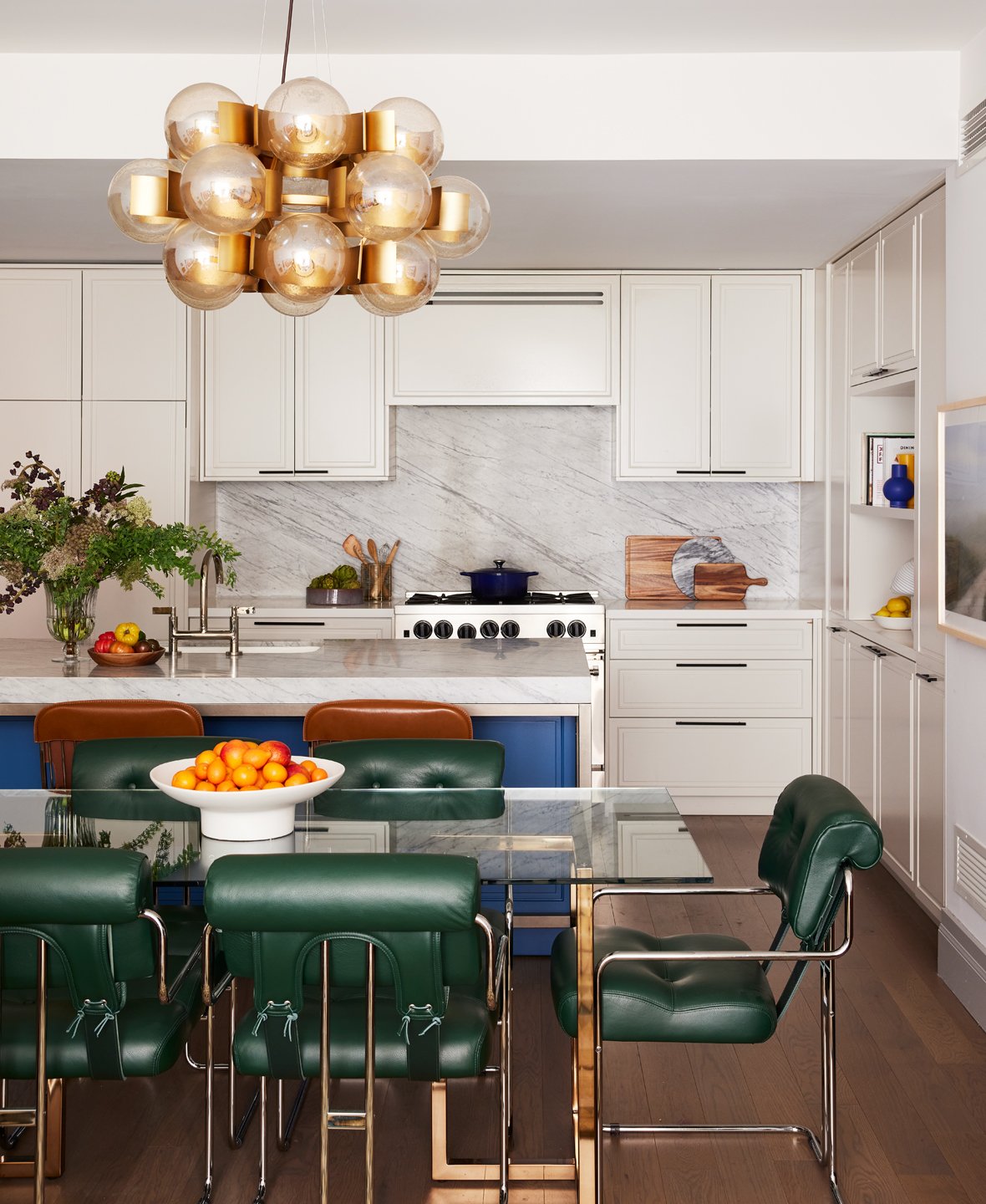 Modern kitchen with white cabinetry, marble backsplash, and a gold and glass spherical chandelier. There is a kitchen island with a bowl of oranges, a dining table with six green leather chairs, and a blue kitchen island. The kitchen has a mix of natural and decorative elements, including vases, a cutting board, and a plant.