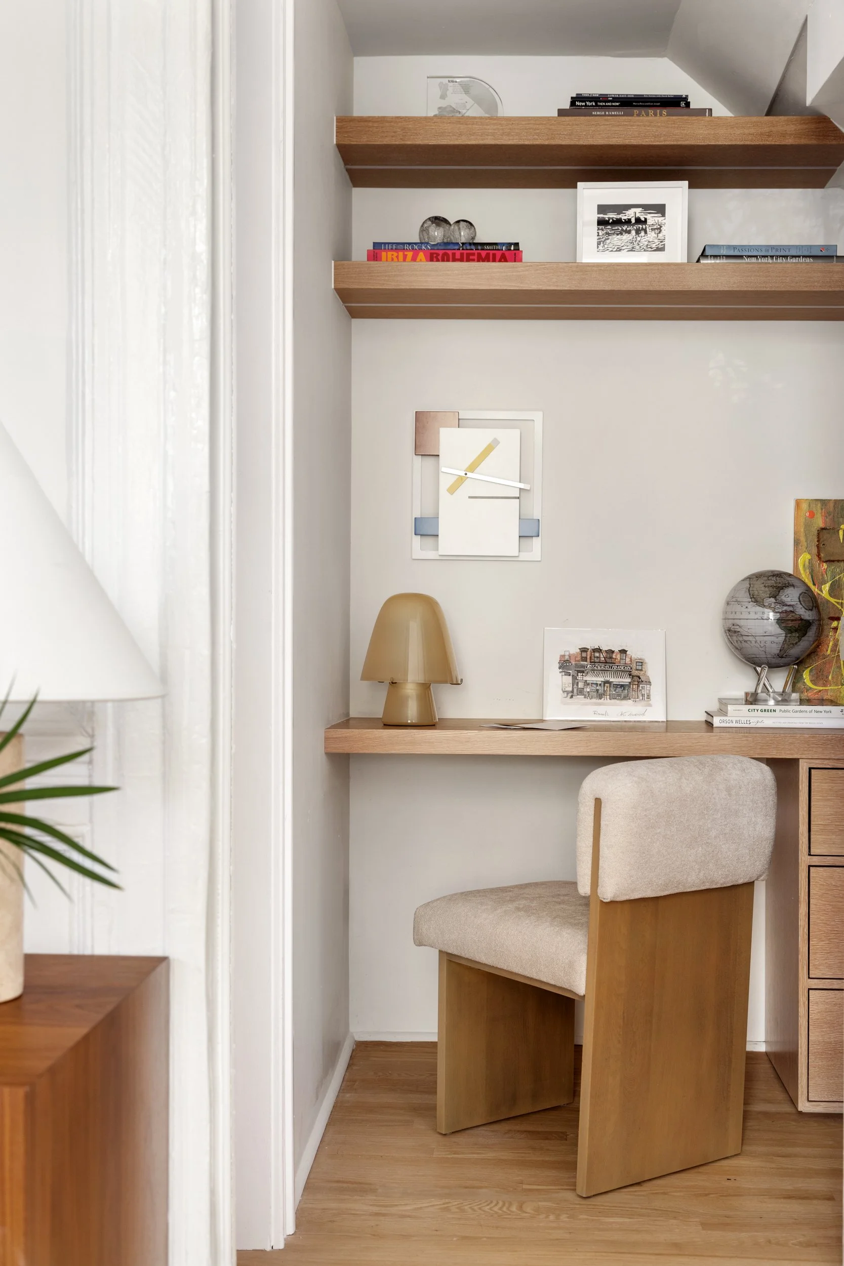 Interior of a home office or study corner with a wooden desk, beige cushioned chair, decorative items, books, and floating shelves on the wall.
