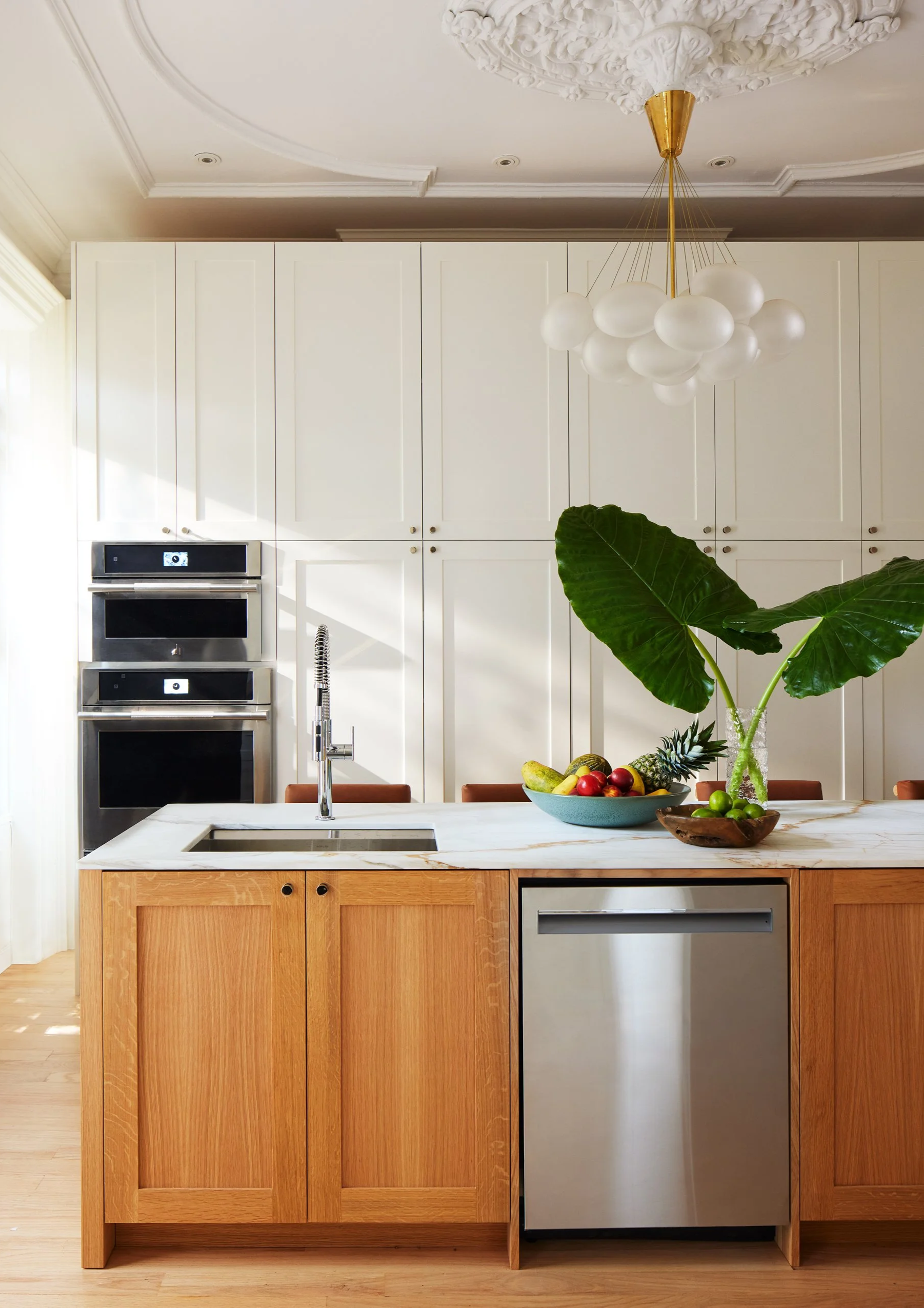 Modern kitchen with white cabinetry, a marble island with wood cabinetry, a fruit bowl, large green plant, built-in ovens, and a contemporary chandelier.