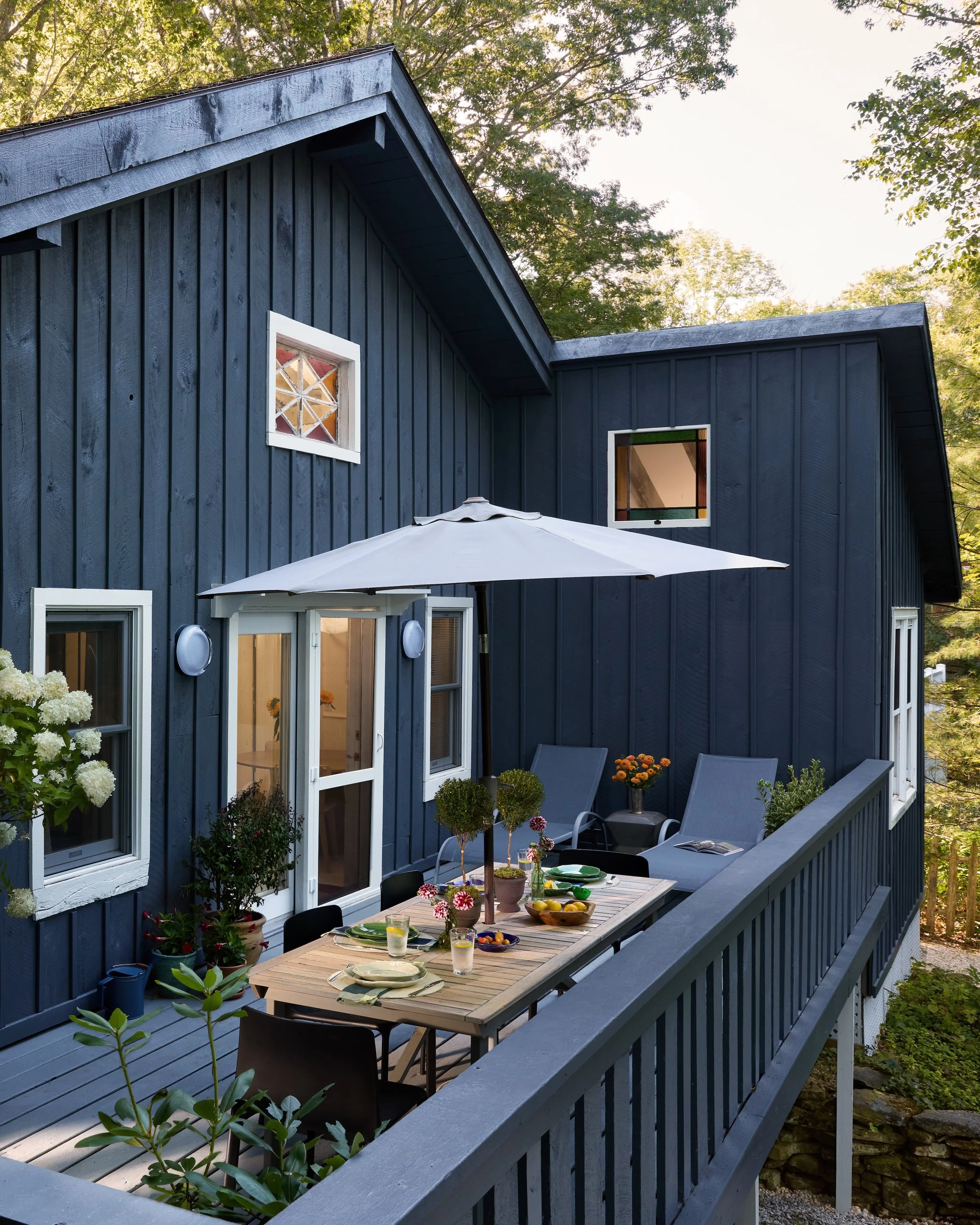 Outdoor patio with dining table, chairs, and umbrella on a balcony attached to a blue house, surrounded by trees and plants.