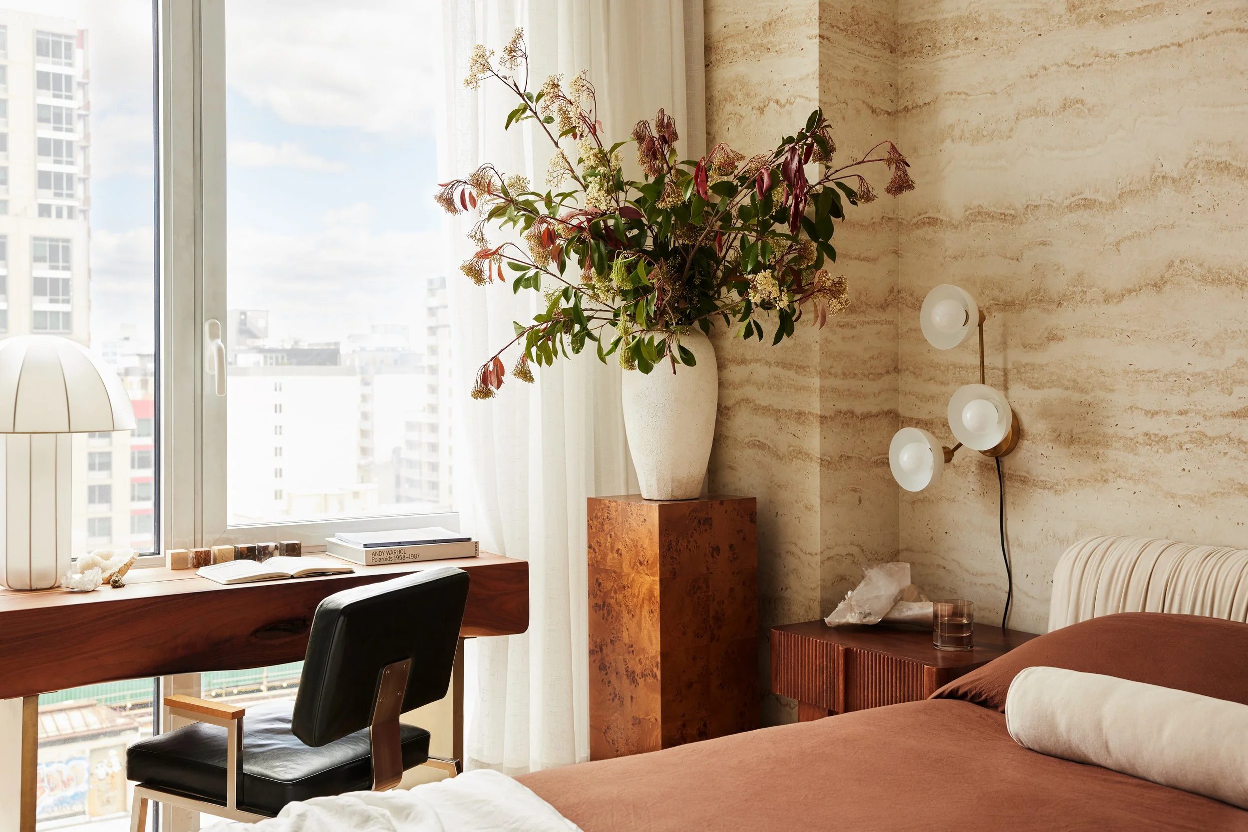 Interior of a modern bedroom with a large window, a desk with a chair, a butterscotch-colored bed with a pillow, a side table with a crystal, and a tall vase with flowers.