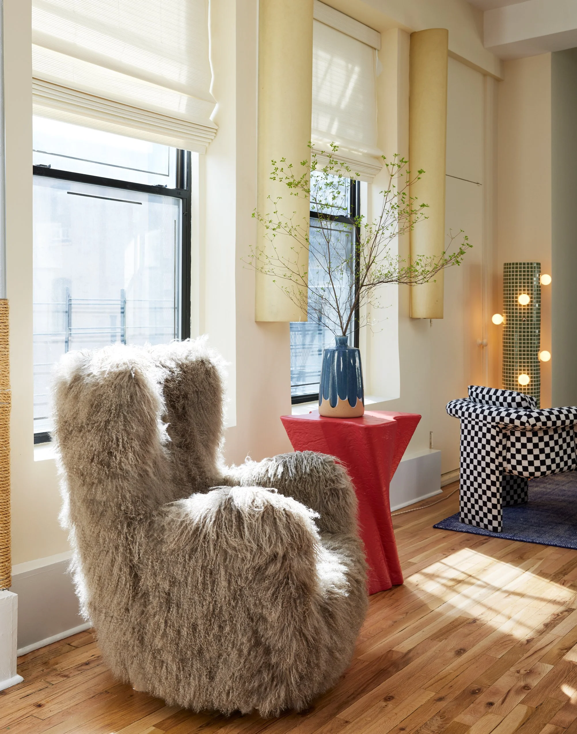 Cozy living room corner with a fluffy beige armchair, a red side table with a blue vase and branches, and a black and white checkered armchair, illuminated by sunlight and decorative string lights.