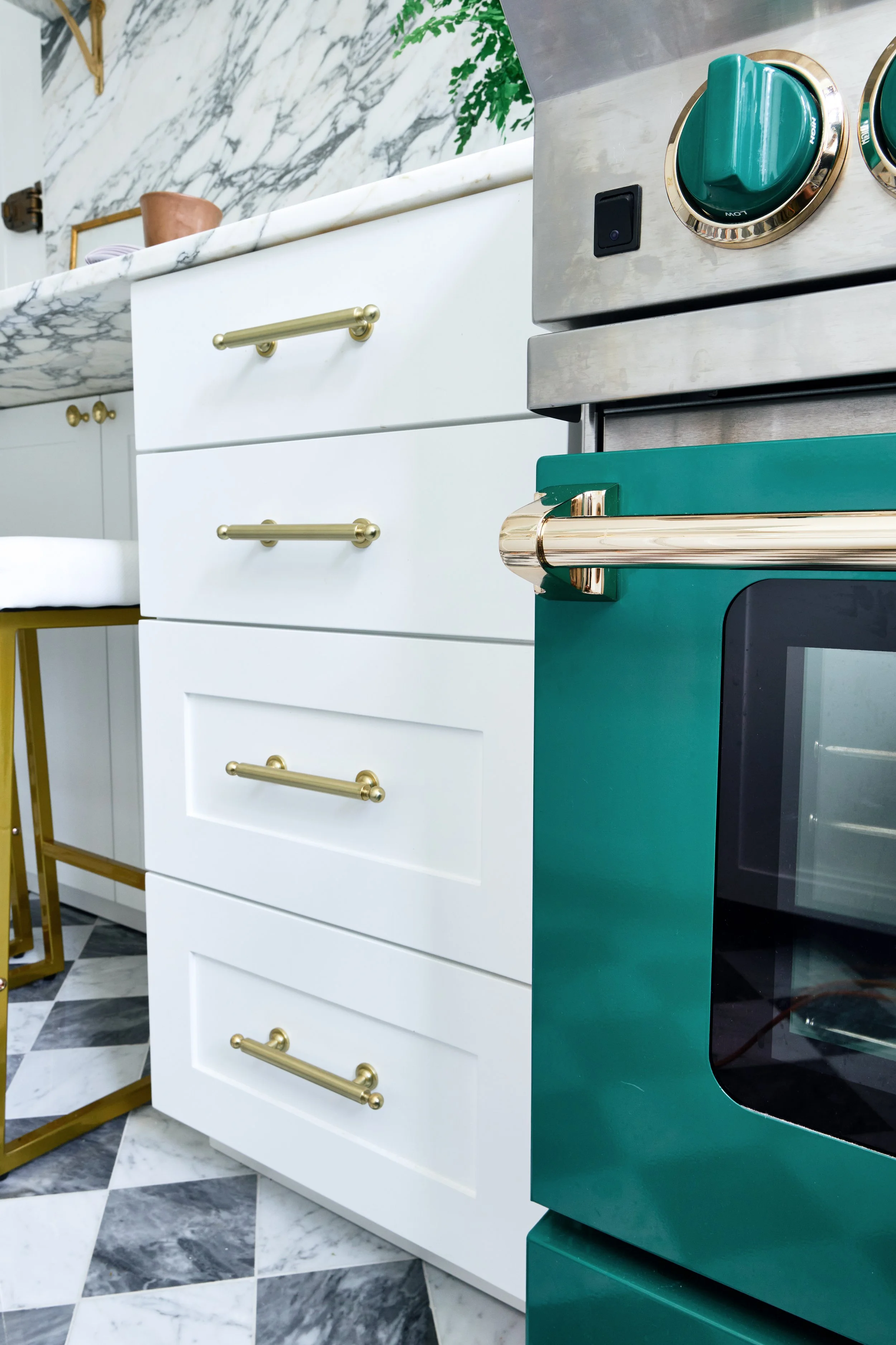 Close-up of a modern kitchen with white cabinets with gold handles, a marble countertop, and a green stove with gold accents. Part of a black and white checkered marble floor is visible.