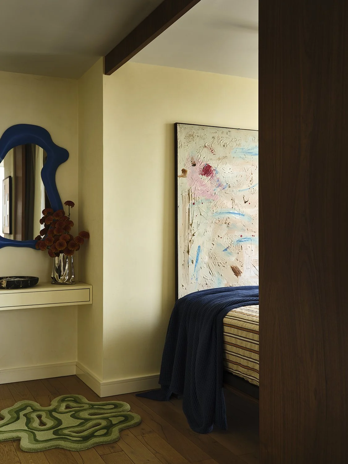 Part of a bedroom with a bed, large abstract painting on the wall, and a small desk with a mirror and vase of flowers.