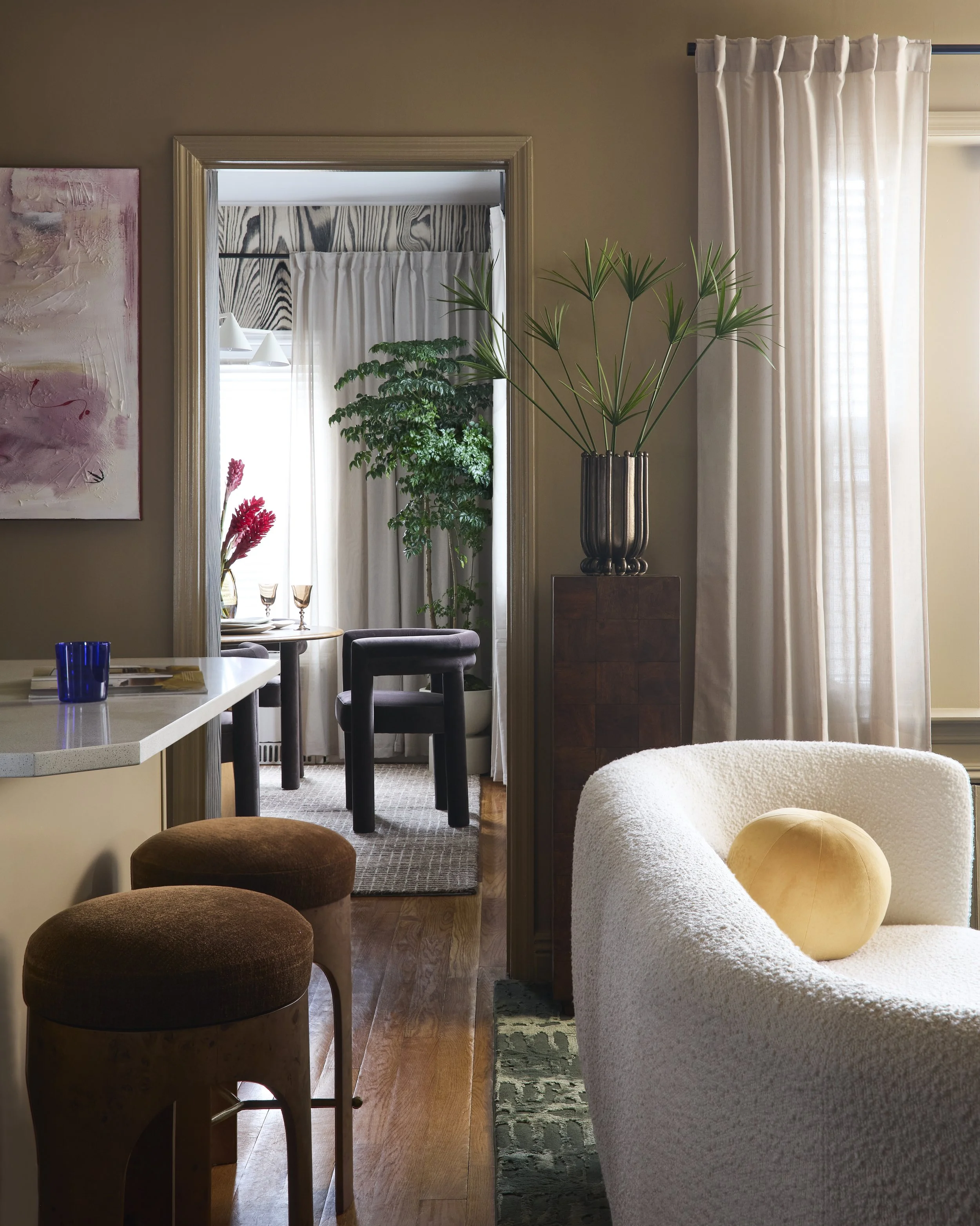 Interior view of a living room with a white curved chair, a yellow round pillow, large window with curtains, a tall potted plant, a wooden side table, and a doorway leading into a dining area with a round table and chairs.