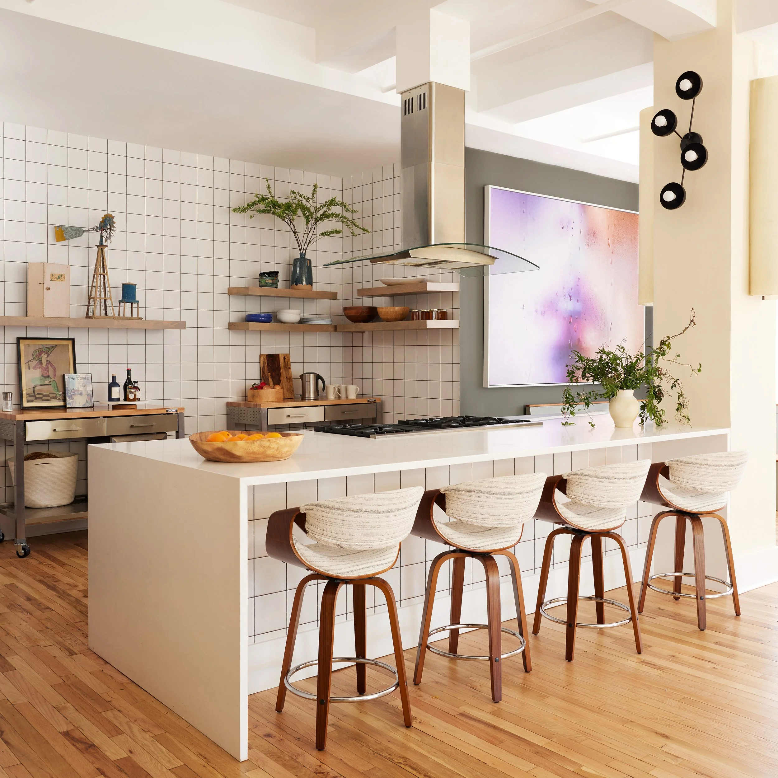 Modern kitchen with white island countertop, bar stools, open shelving with decor items, and a large abstract painting on the gray wall.