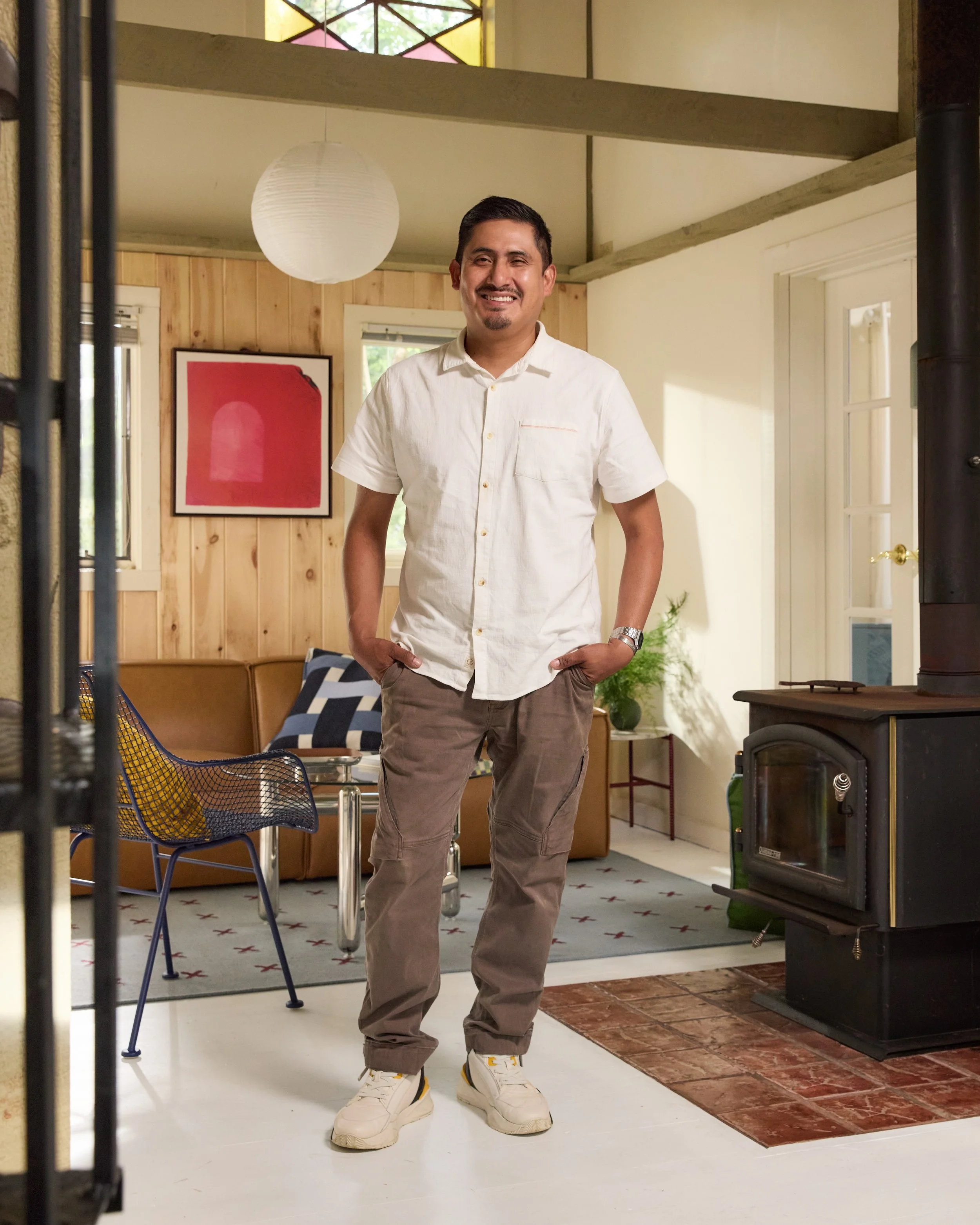 A man standing in a cozy living room, smiling, with white shirt, brown pants, and sneakers, in front of a leather couch, artwork on the wall, and a wood stove.