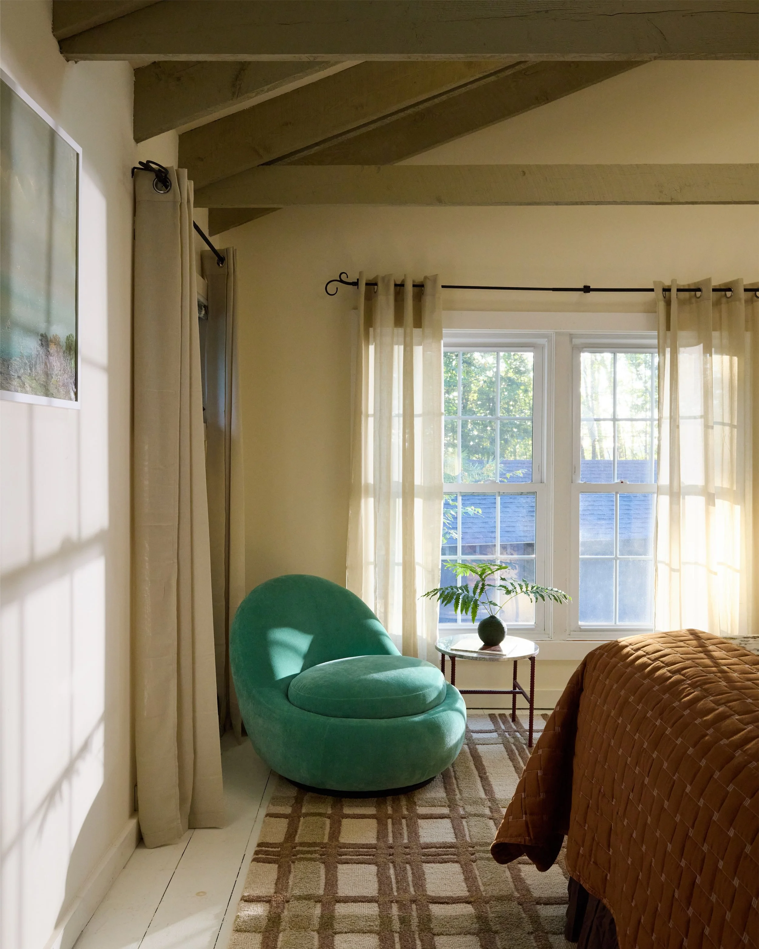 A cozy bedroom corner with a green upholstered chair, a small round table with a potted plant, window with sheer cream curtains, and part of a bed with a brown quilt.