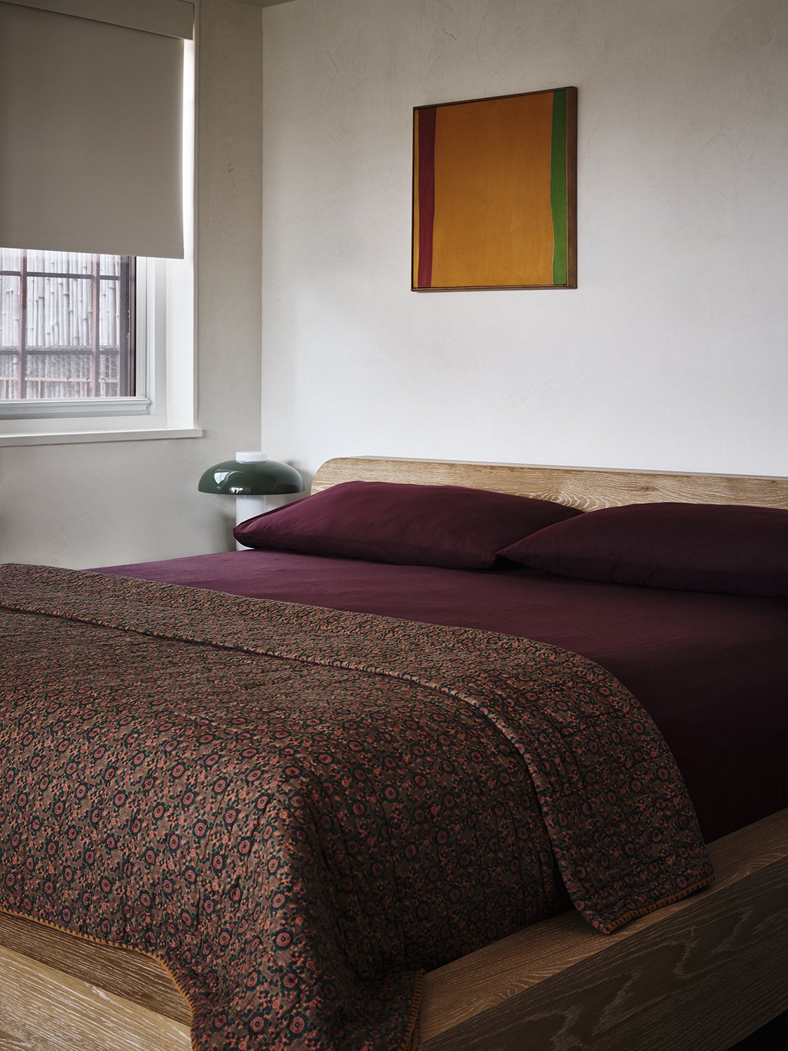A bedroom with a bed covered in a dark red bedspread and matching pillows. The bed has a wooden headboard, and there is a small window with a blind partially open, showing a wooden fence outside. A green and white lamp is beside the bed, and a colorful abstract painting hangs on the white wall above the bed.