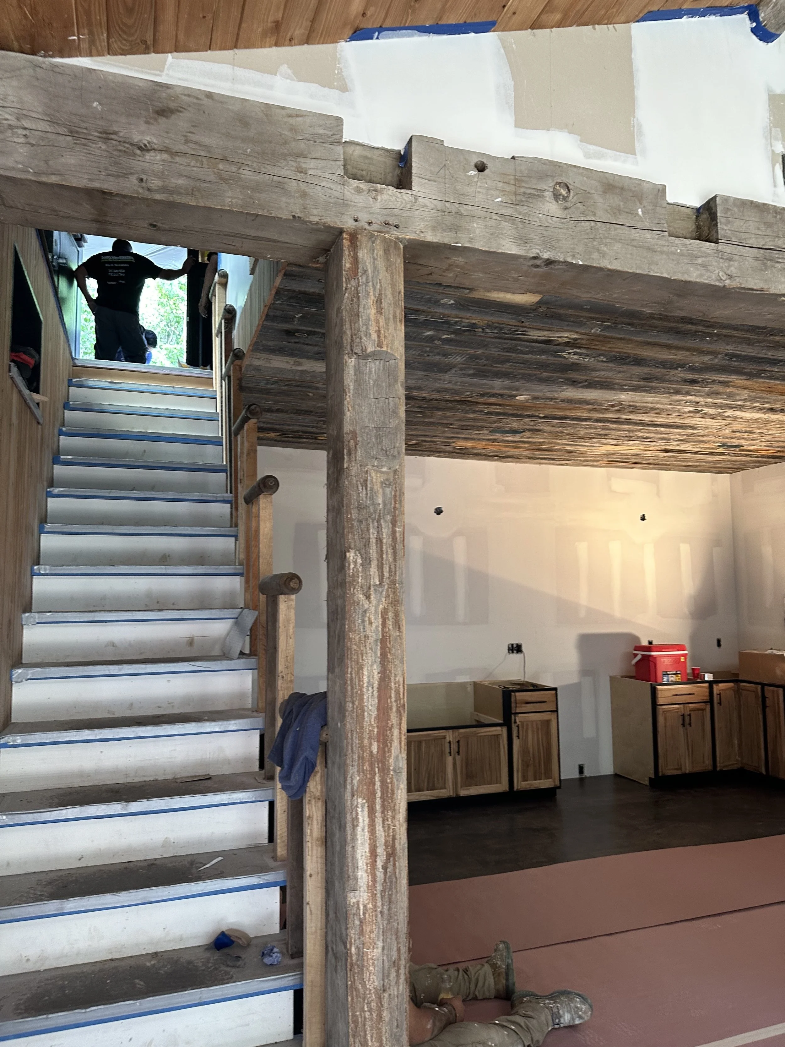Construction site interior showing unfinished wooden stairs, a partially completed second floor, and a person lying on the floor. Experts and tools are visible in the background.