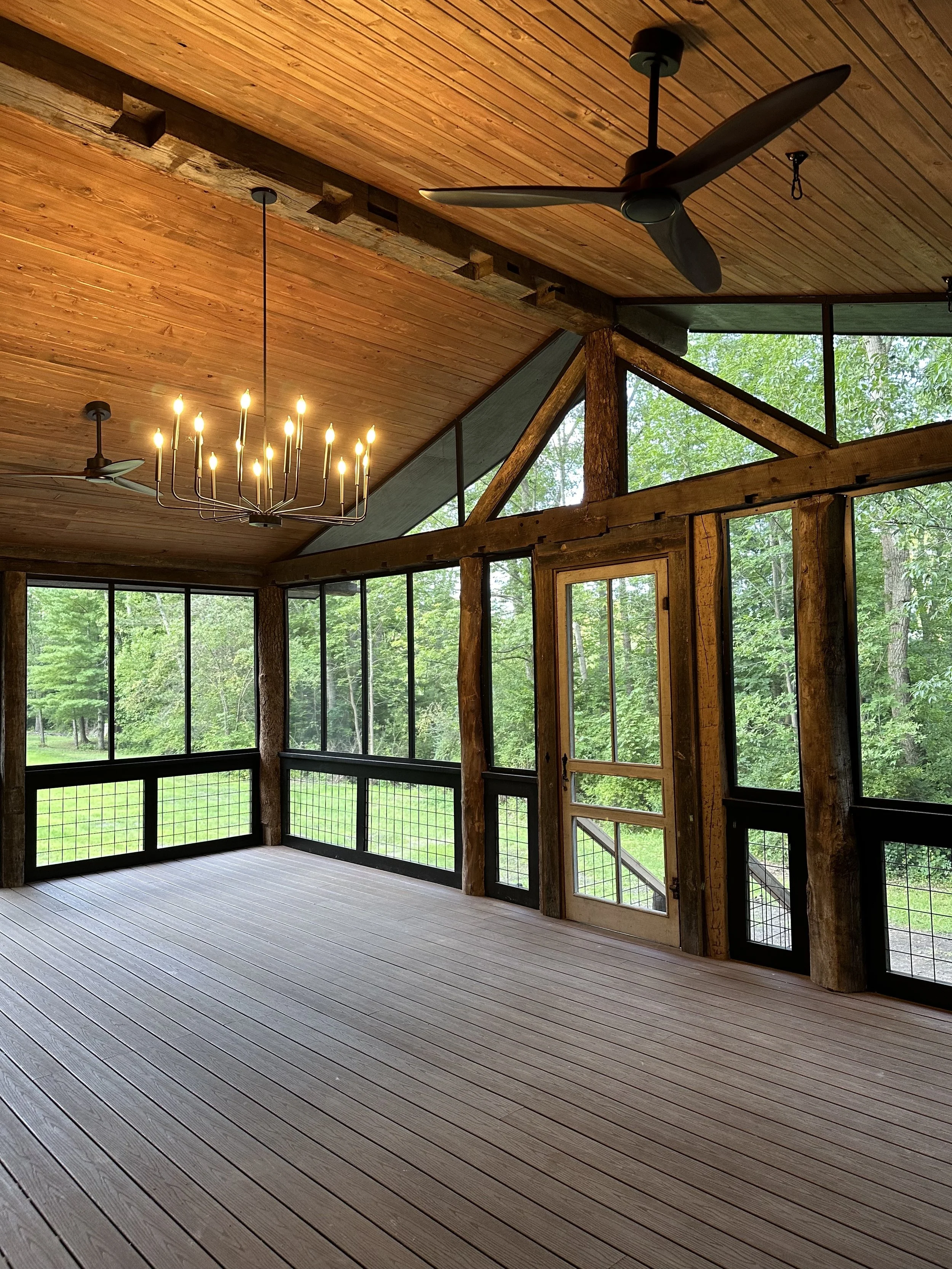 A spacious screened-in porch with wooden floors, wooden ceiling, and large glass windows showing green trees outside. There is a chandelier and ceiling fans.