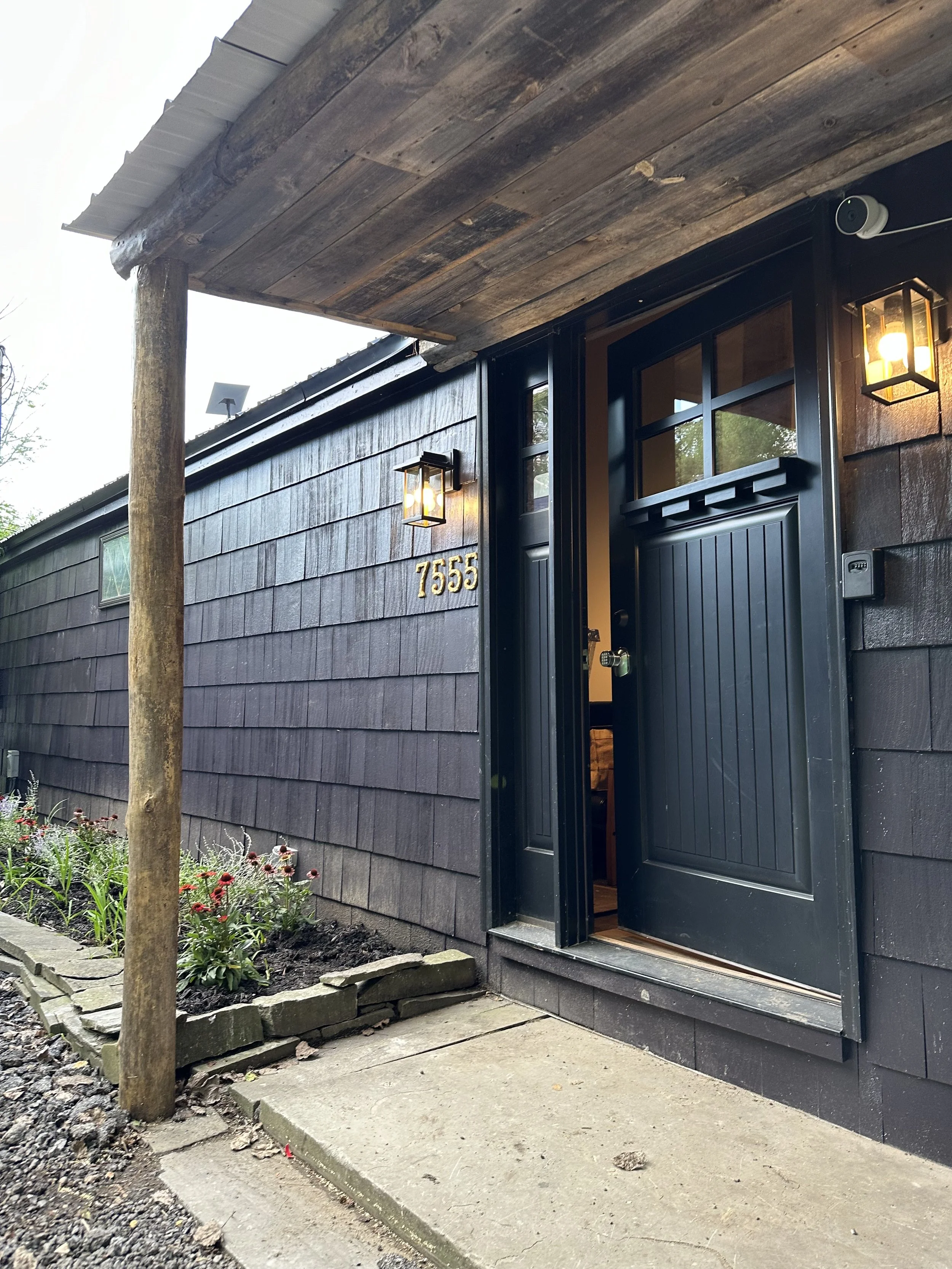 Close-up of the entrance to a house with a black door, the house has dark wooden siding, with the number 7555 on the wall, and outdoor lighting fixtures illuminating the doorway.