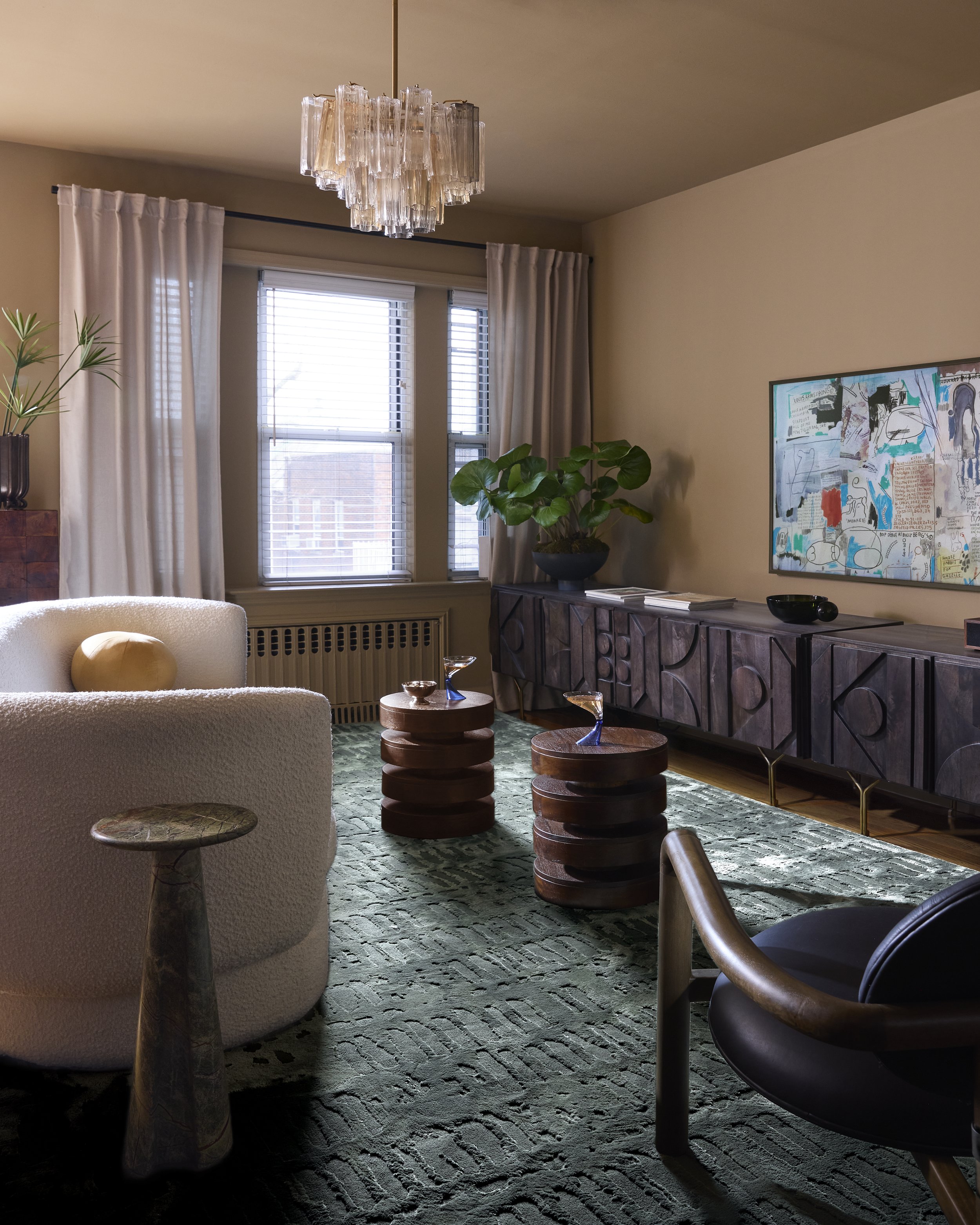 Living room with beige walls, a large window with blinds and cream curtains, a chandelier, a white curved sofa with a yellow pillow, two wooden side tables with glass top decorations, a dark wood sideboard with large plant, artwork on wall, and a patterned green carpet.