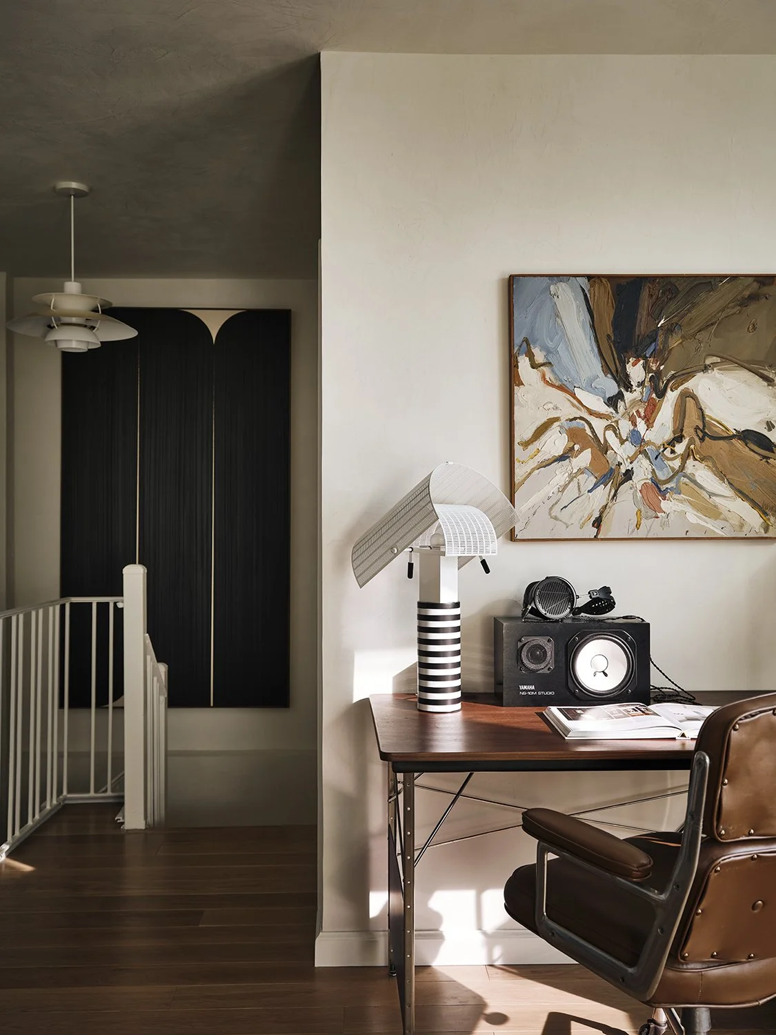 Interior view of a workspace with a wooden desk, a brown leather chair, a white and black striped lamp, a speaker, and an abstract painting on the wall.