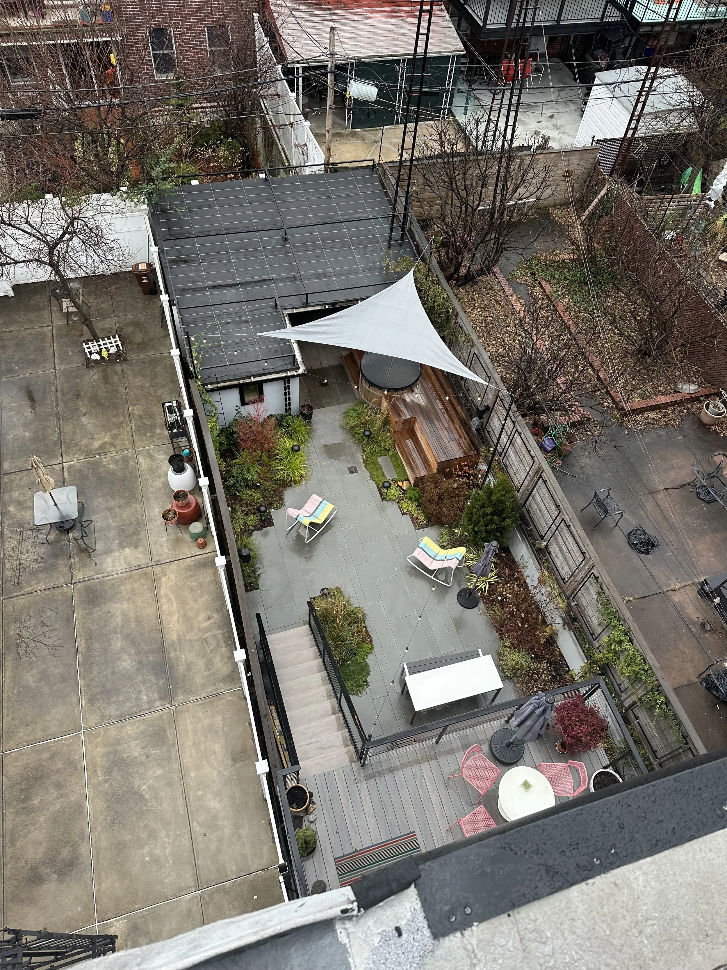 An aerial view of a rooftop patio with outdoor furniture, potted plants, a shade sail, and a surrounding fenced garden area. There is a adjacent concrete space with a small table and chairs.