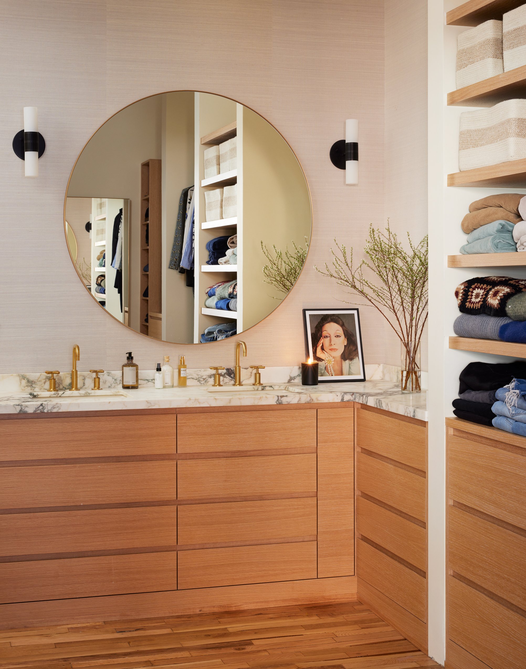 Modern bathroom with wooden cabinetry, marble countertop, round mirror, and open shelving filled with folded towels and clothes. Decorative items include a framed photo, candle, and plant accents.