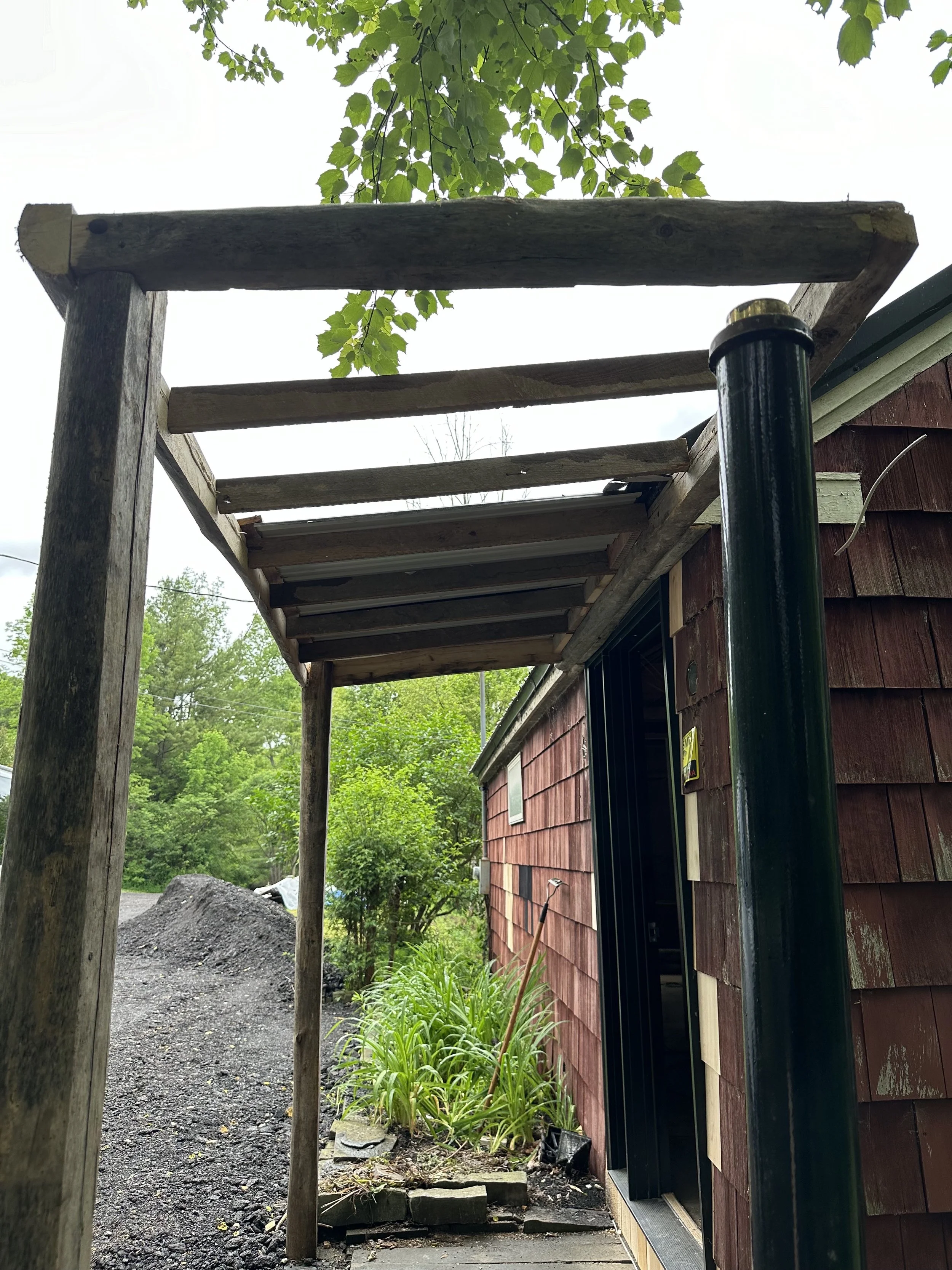 Construction of a wooden porch or deck extension with support posts next to a red house with shingles, greenery, and overcast sky in the background.