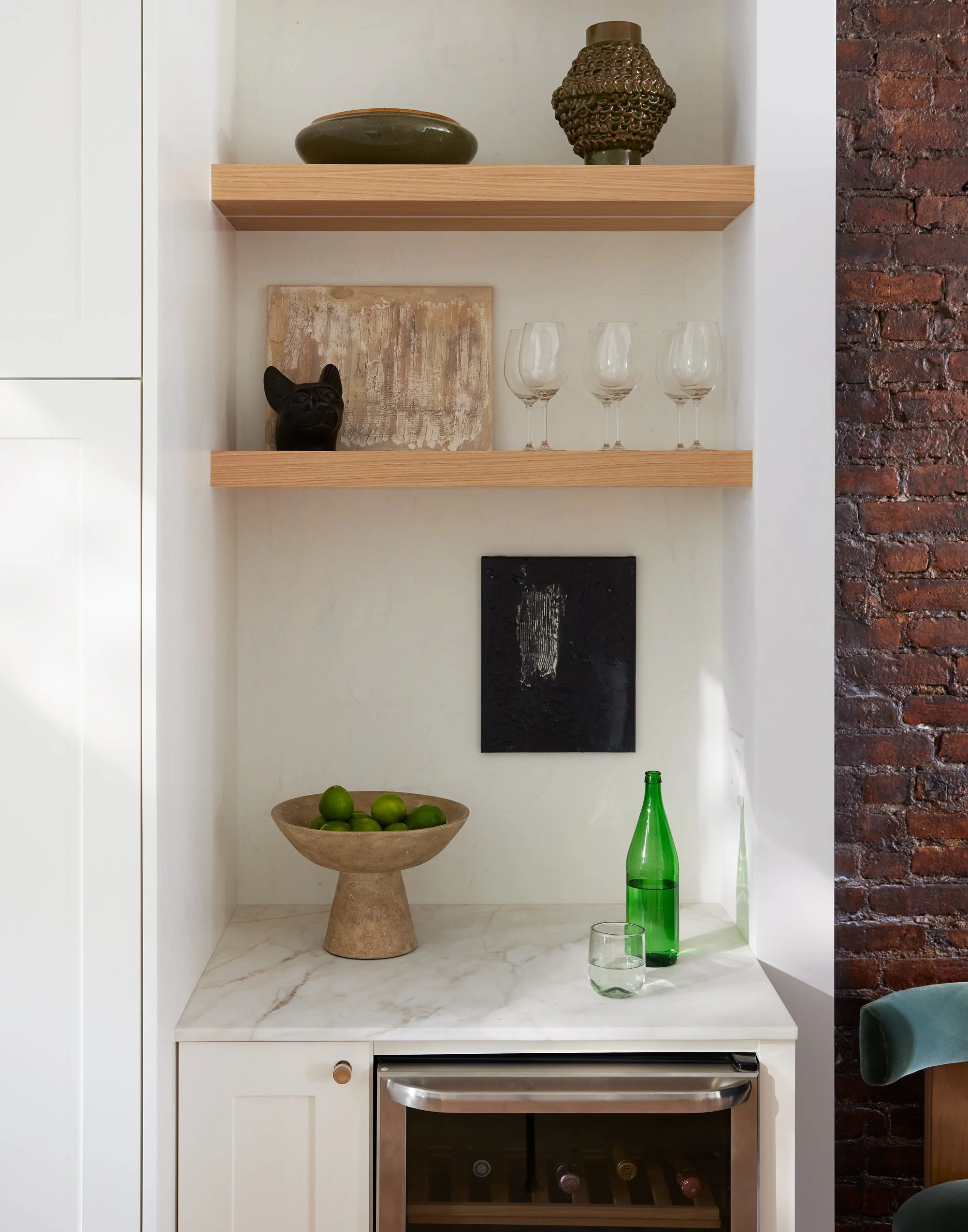 Kitchen nook with white cabinetry, wooden shelves holding decorative objects, a marble countertop with green limes in a stone bowl, green glass bottle and glass, and wall art, against a brick wall.