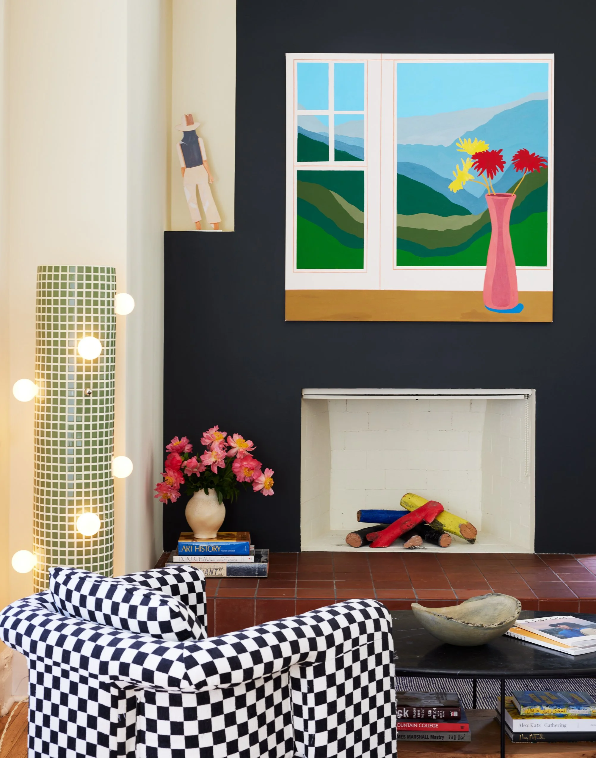 Living room with a checkered black and white armchair, a black coffee table with books and a ceramic bowl, a white fireplace with colorful logs, a pink flower vase on a small side table, a large painting of mountains and a window scene on the dark wall, a vase with pink flowers, a tall lamp with green square patterns, and a wooden figure on a shelf.