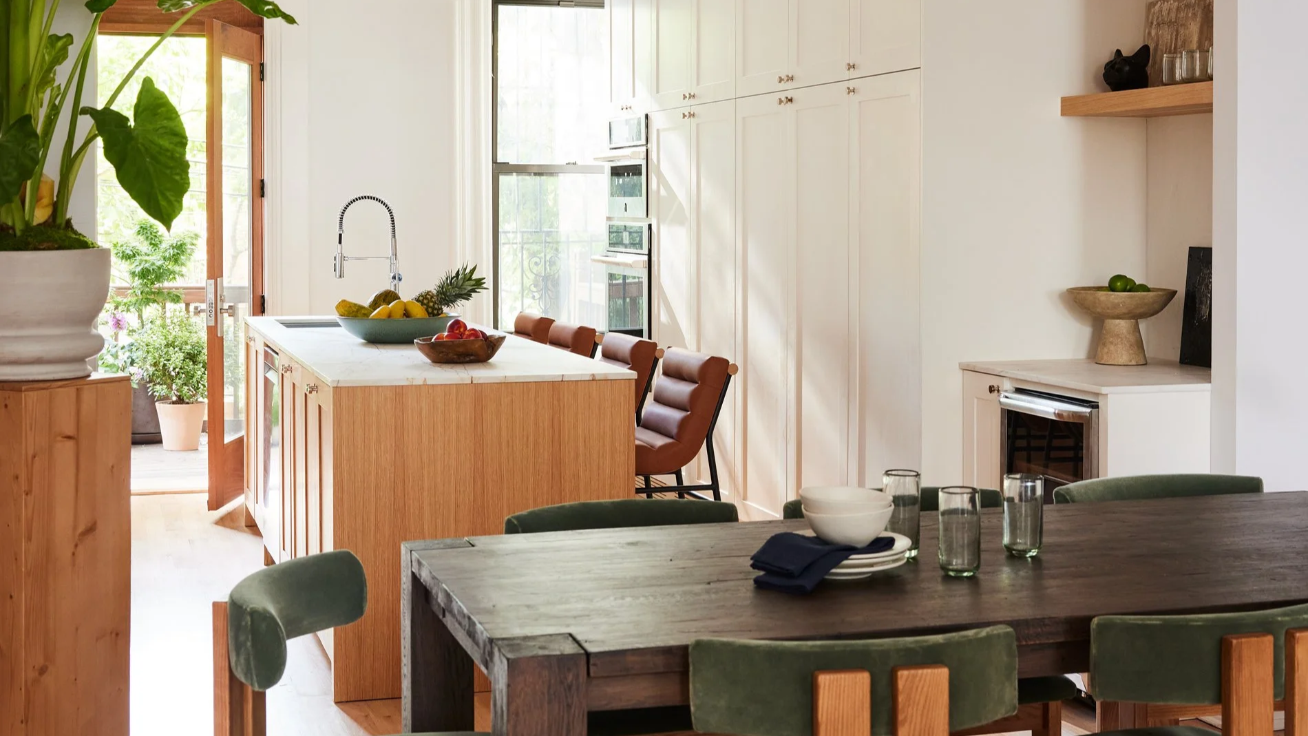 Kitchen with a wooden island, fruit bowl, and brown chairs in front of large windows, with a dining table set with bowls and glasses in the foreground.
