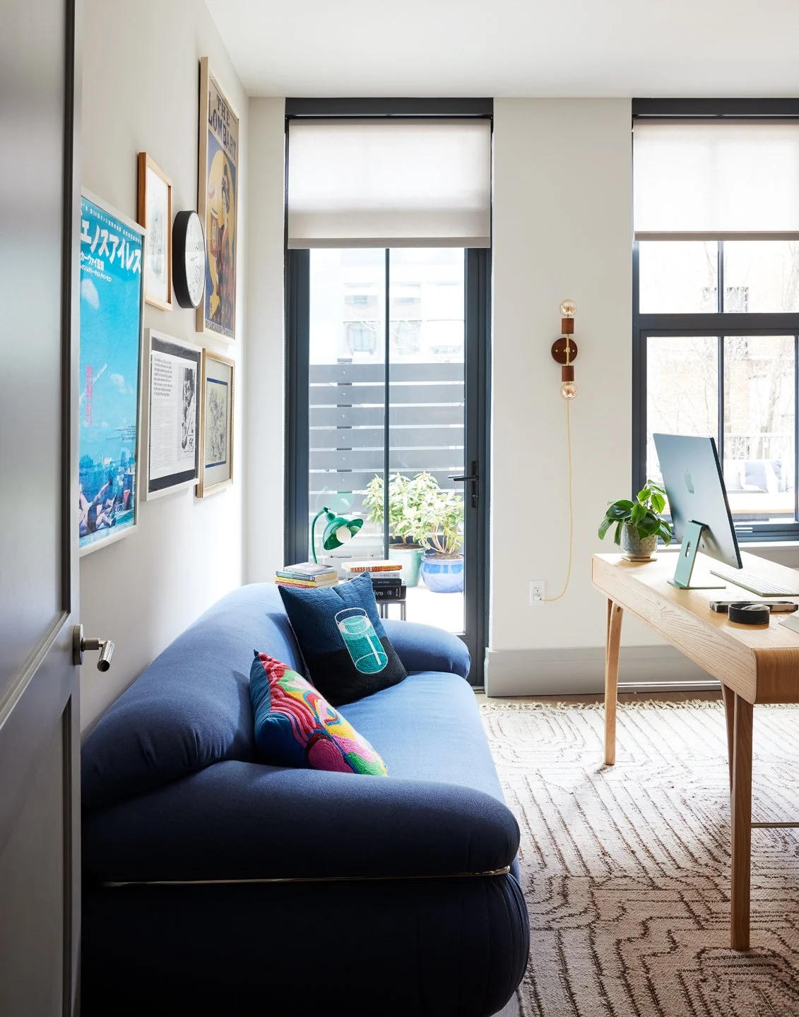 A modern living room with a blue sofa, colorful pillows, wall art, a wooden desk with a computer, a potted plant, and a sliding glass door leading to a balcony with outdoor plants.