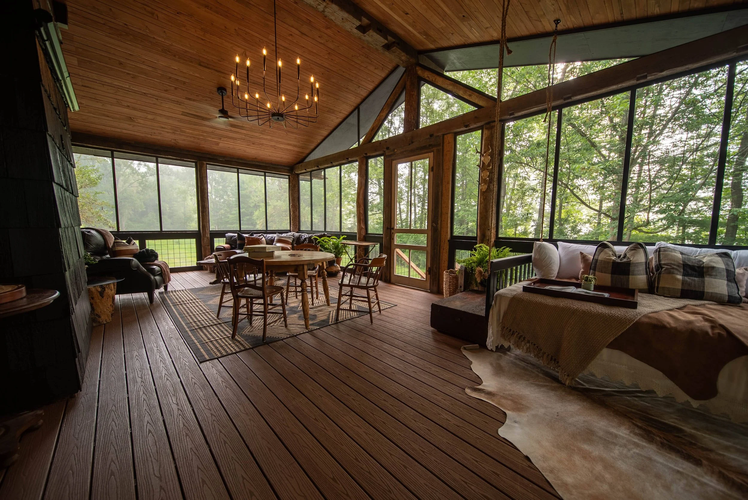 A rustic sunroom with wooden floors and ceiling, large windows showing green trees outside, a chandelier hanging from the ceiling, a round dining table with chairs, leather armchairs, a patterned rug, and a cozy sitting area with a couch and throw pillows.